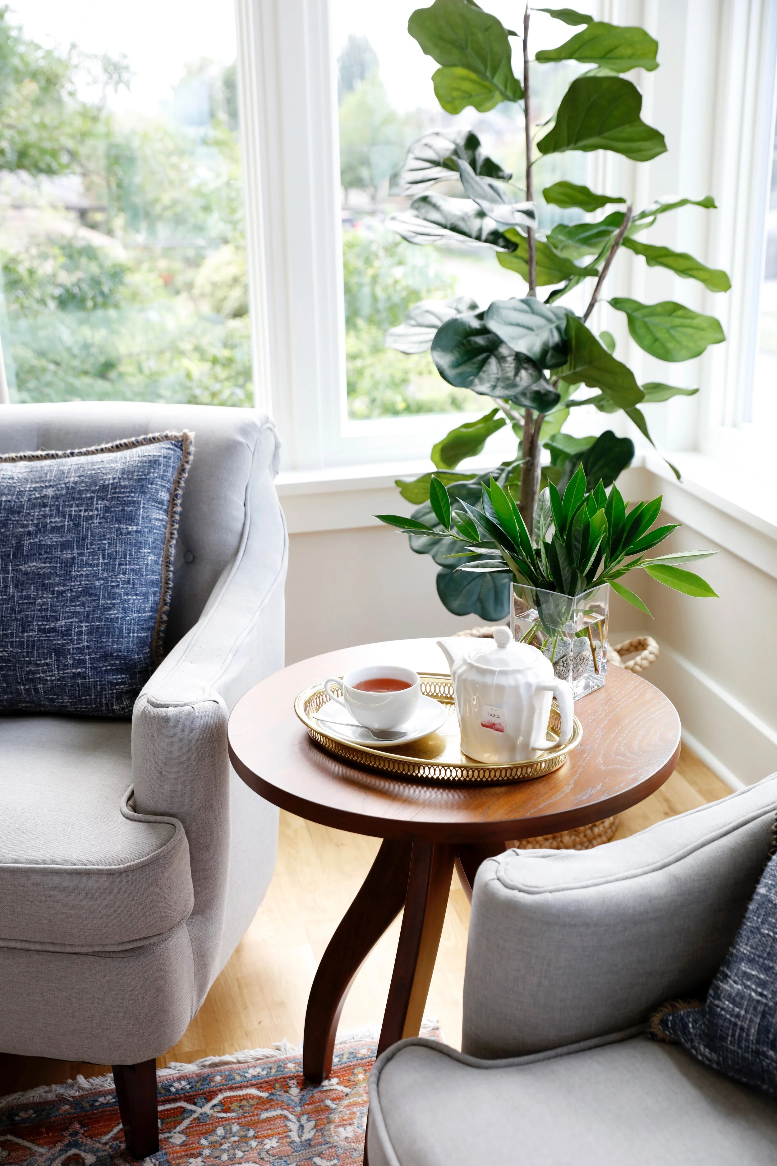 A cozy living room corner featuring two light gray armchairs with dark blue cushions, a small round wooden table with a tea set and a potted plant, large windows with white frames allowing natural light, and a leafy green indoor plant in a glass container.