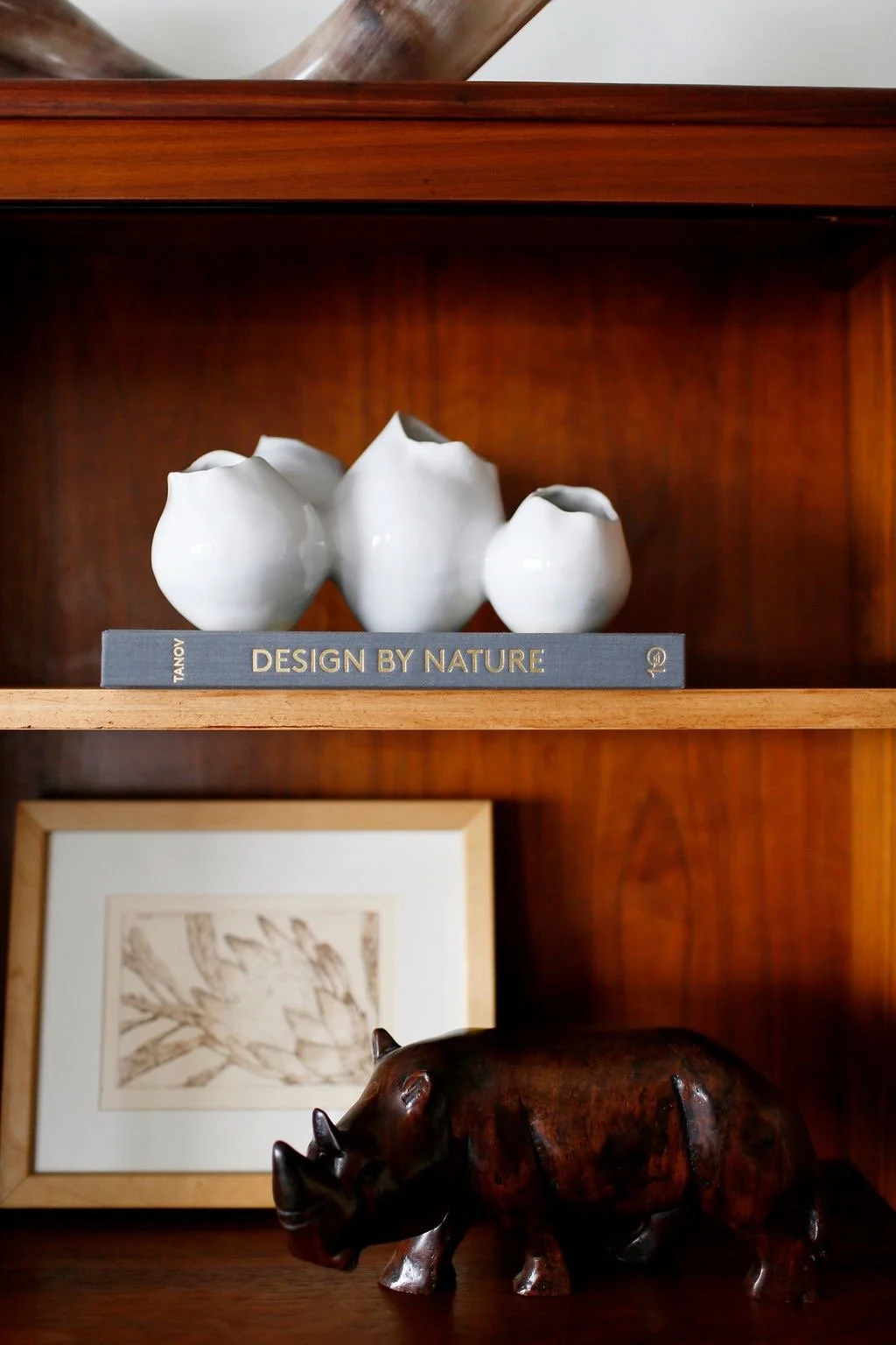 Three white ceramic vases of varying sizes on a gray book titled "Design by Nature," with framed artwork of abstract plant-like design and a wooden hippopotamus figurine on a wooden shelf.
