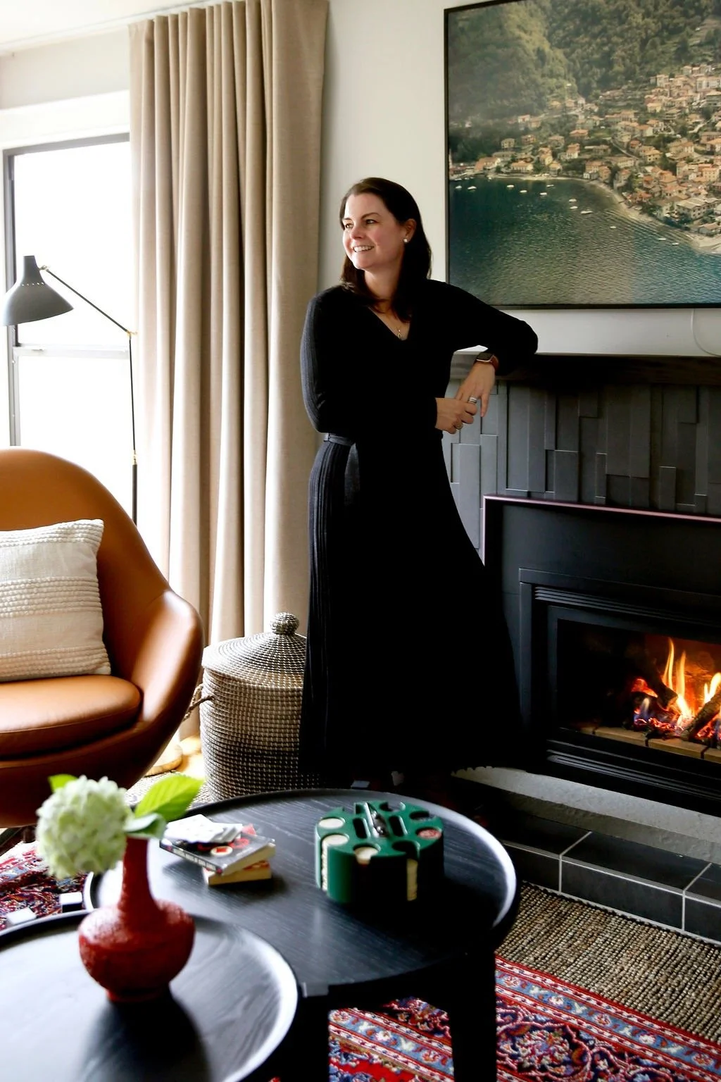 A woman in black standing near a fireplace in a cozy living room, with a large painting of a coastal landscape behind her, a leather armchair with a cushion, a small table with a flower vase, and a rug.