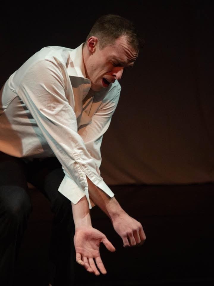 A man wearing a white dress shirt is crying and holding his head in despair, sitting against a dark background.