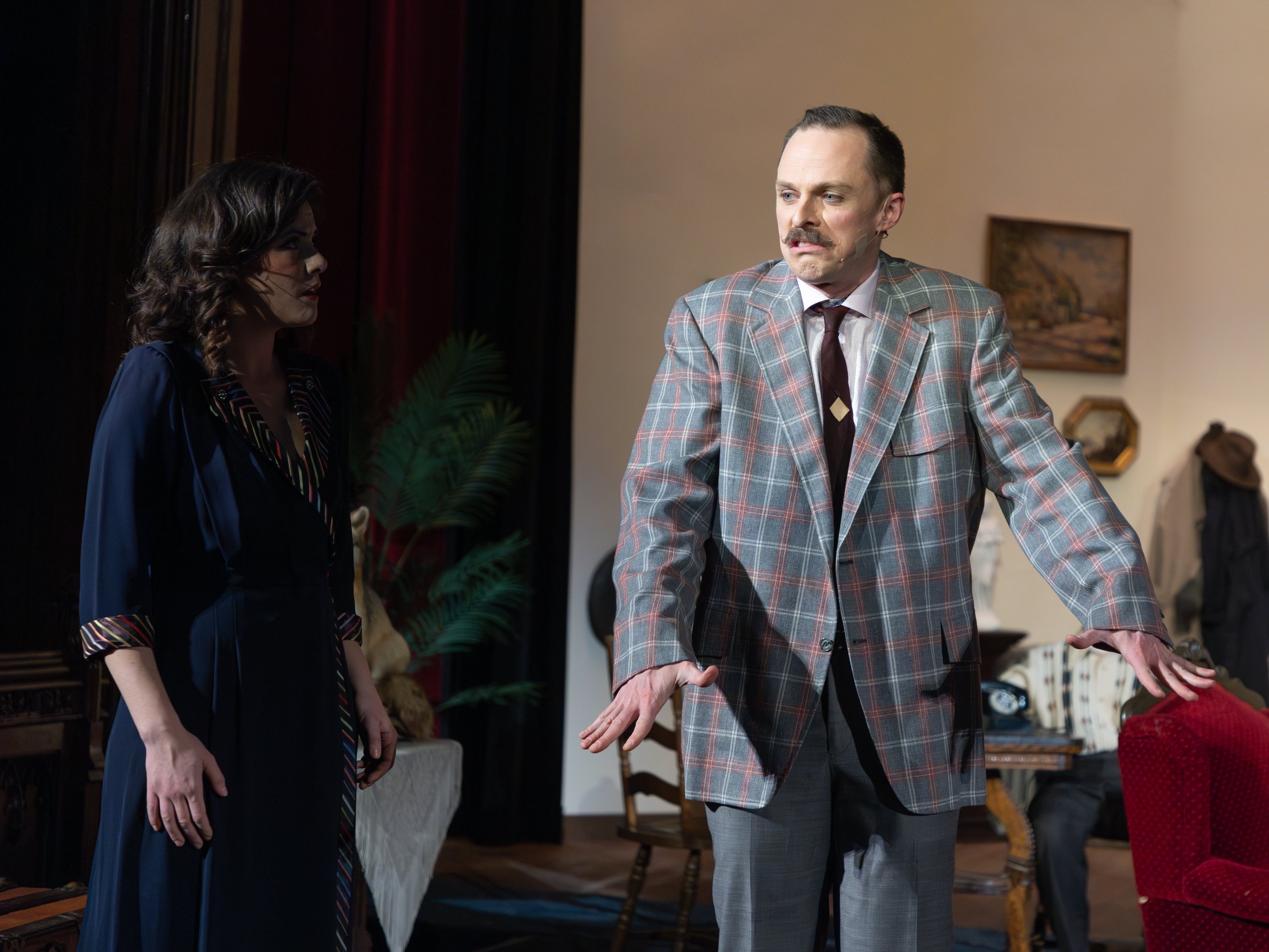 A man in a plaid suit jacket and a woman in a dark dress engaged in an intense conversation in a vintage-style living room.