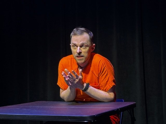 Marcus Sharpe from Another Medea by Aaron Marks is a man wearing an orange prison jumpsuit sitting at a table with a black backdrop, gesturing with his hands.