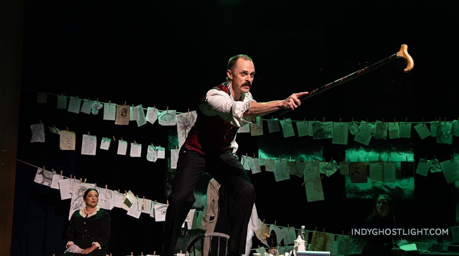 friedrich nietzsche, the leading character in Seeking Nietzsche by Marcia Epic-Harris, is a man with a mustache wearing a white shirt and maroon vest, is performing on stage with a cane. Behind him are illustrations and notes hanging on strings.