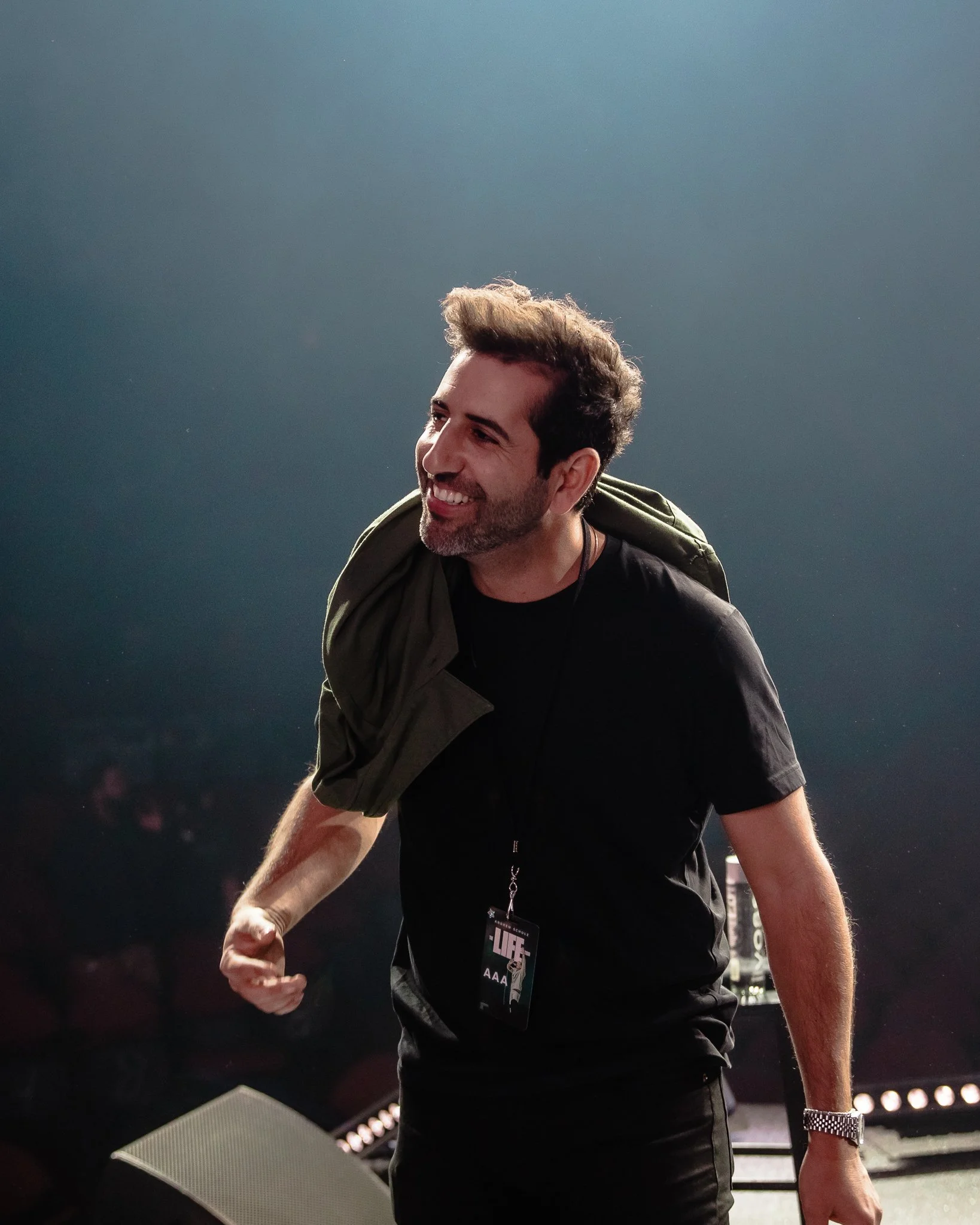 A man smiling and looking to the side, wearing a black t-shirt and a wristwatch, with a large badge hanging around his neck, standing outdoors at night with a dark background.