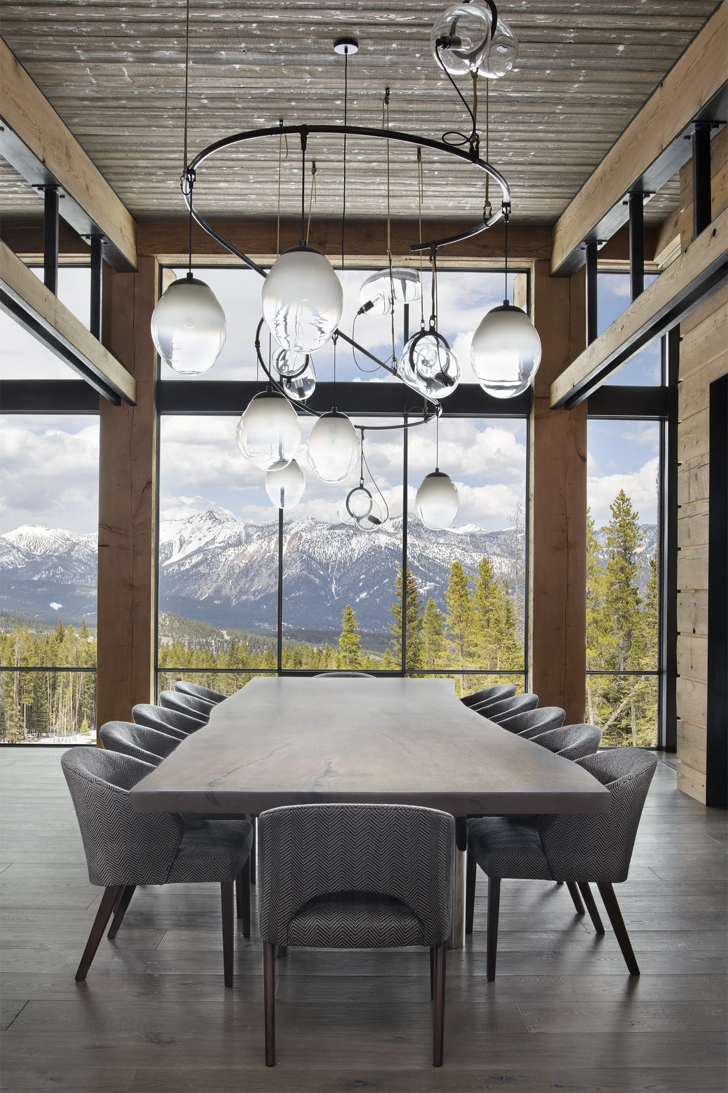 Modern dining room with a large marble table, 10 upholstered chairs, wooden beams, floor-to-ceiling windows with mountain and forest view, and TwentyTwo Brightbound studio-blown architectural lighting fixture.