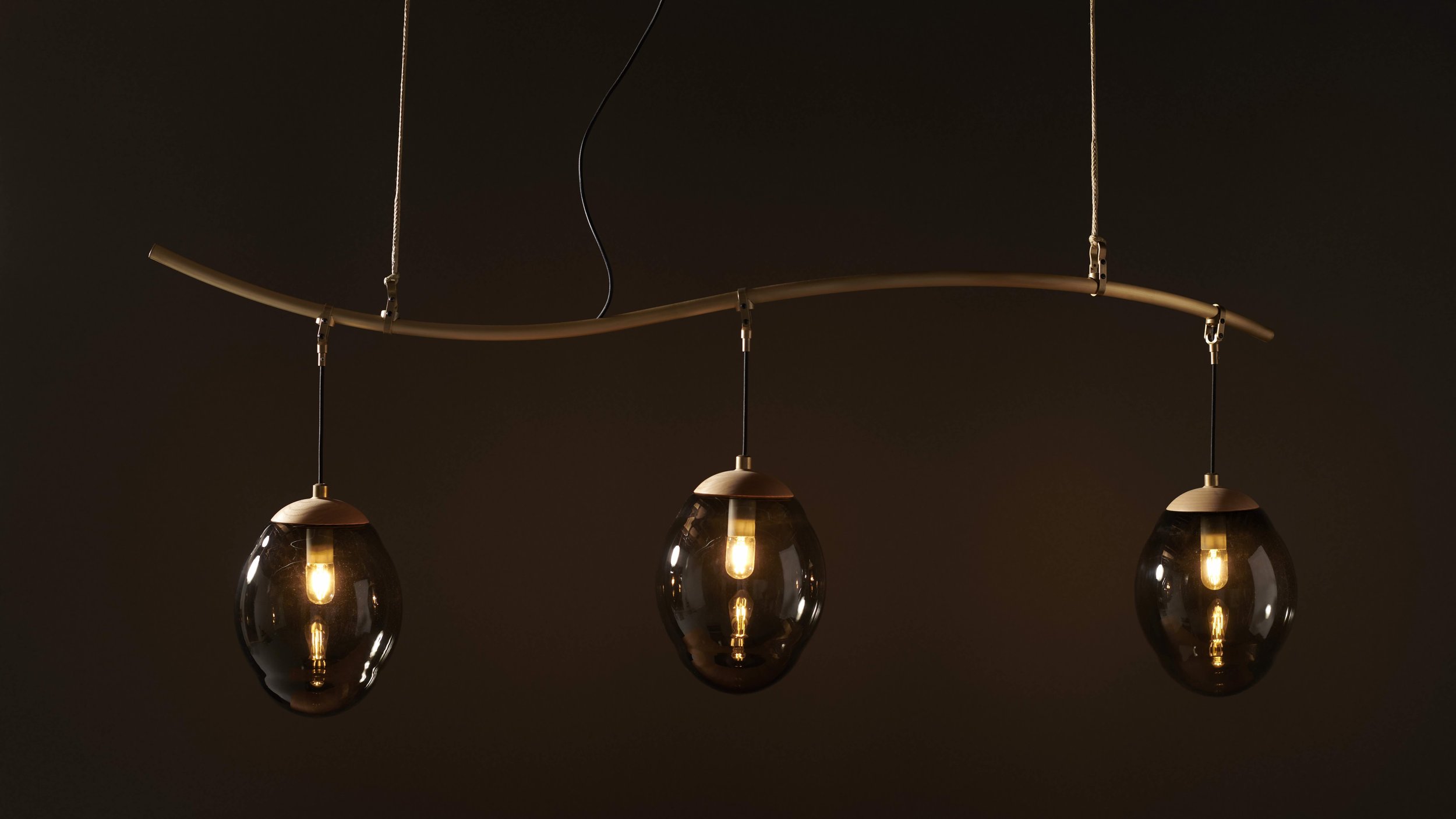 Modern pendant chandelier with three glass bulbs hanging from a curved metal fixture against a dark background.