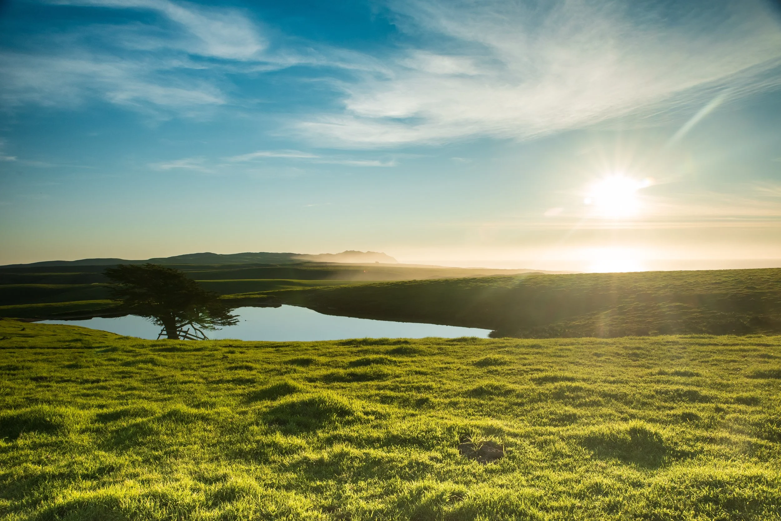 Scenic landscape with a grassy field, a pond, a tree, rolling hills in the distance, and the sun shining in a mostly clear sky with some scattered clouds.