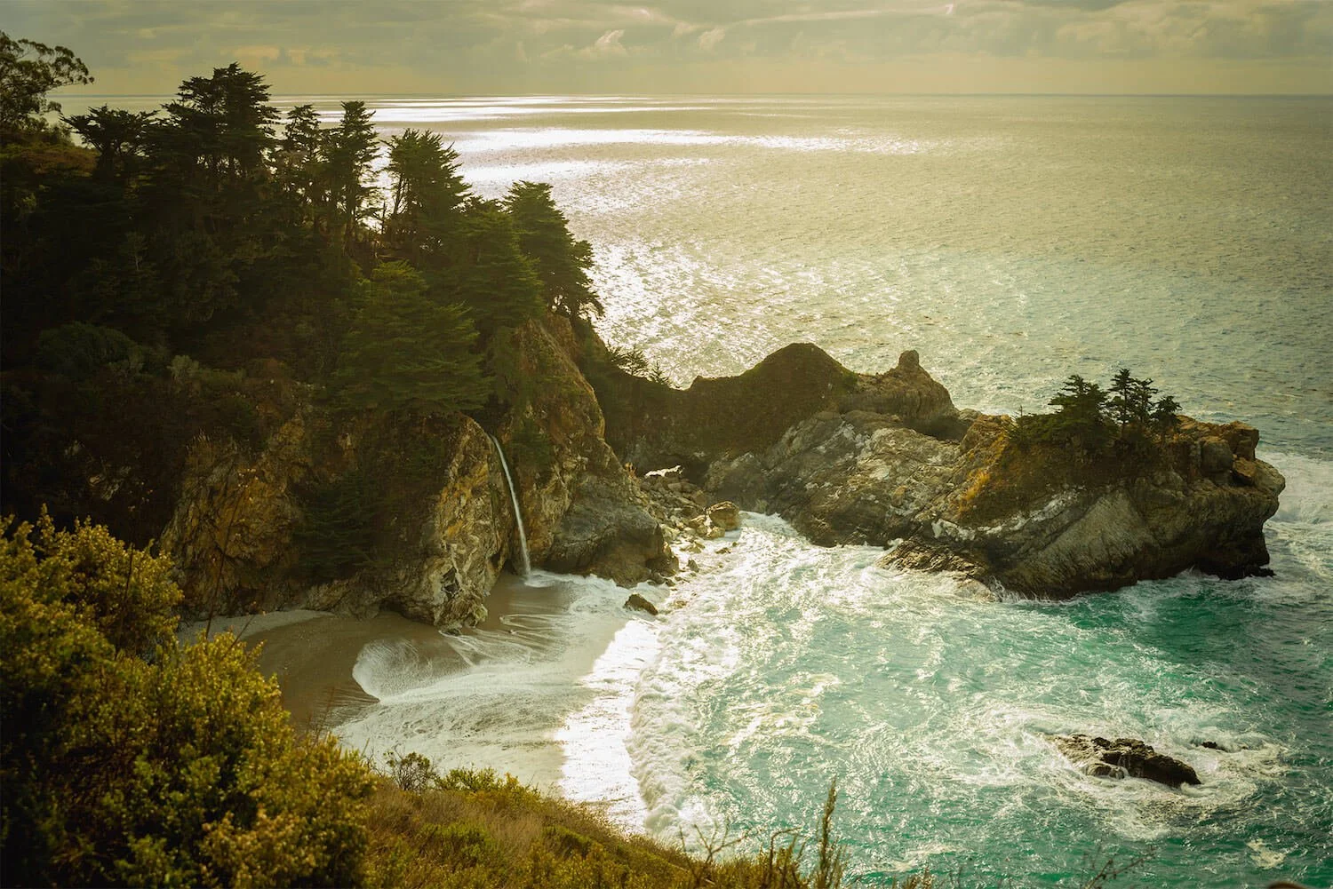 A coastal landscape featuring rocky cliffs covered with trees, a small waterfall flowing into the ocean, and a sandy beach below. The ocean waves are crashing against the rocks, with the sunlight reflecting off the water.