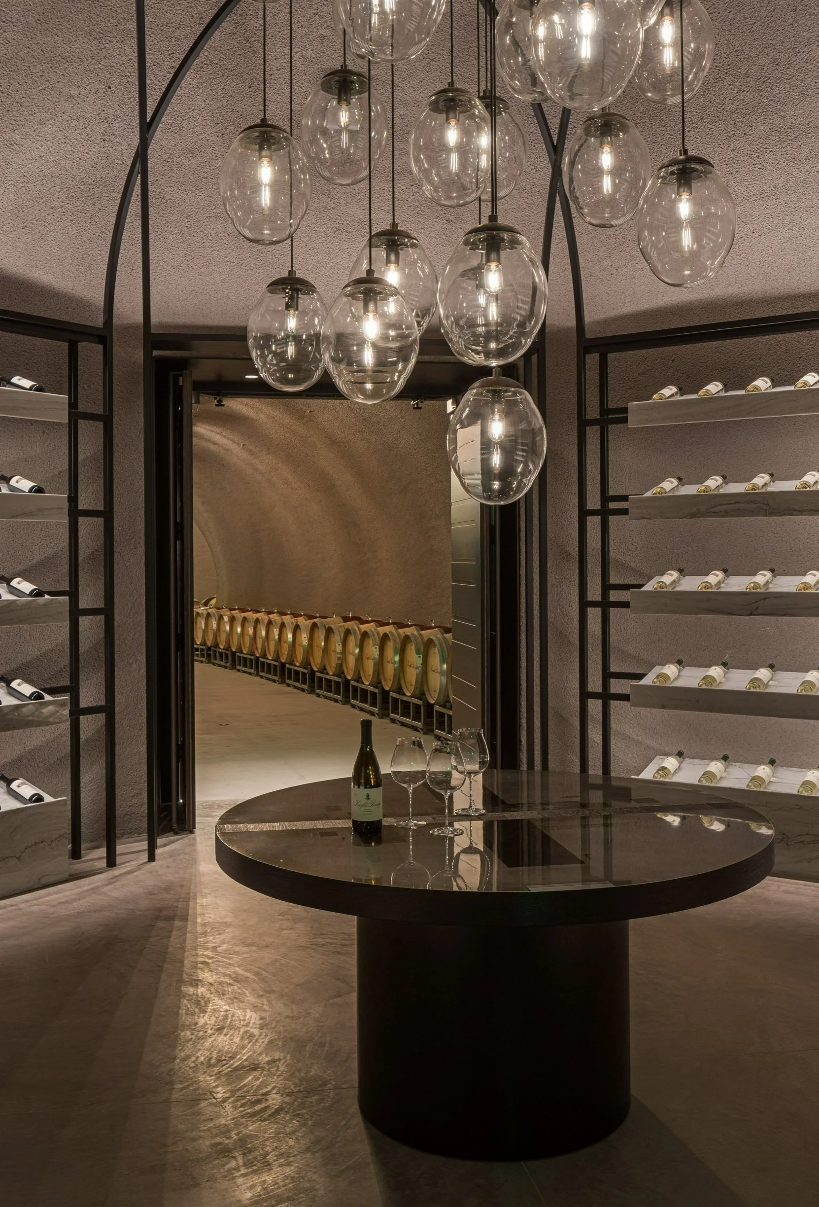Elegant wine cellar with Gemma studio-blown glass lighting chandelier, a round reflective table in the center, and shelves filled with wine bottles on either side, with a seating area visible through a doorway in the background.