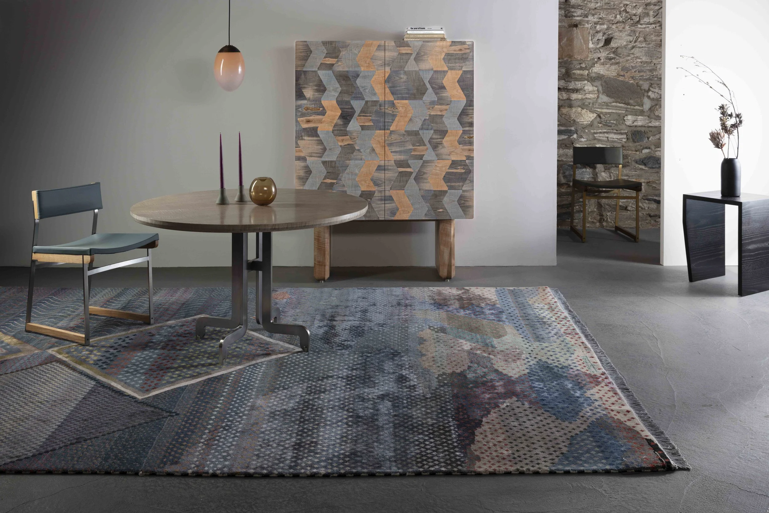 Modern living room with abstract rug, wooden table with two candles and a vase, patterned artwork on the wall, chair, and black side table with a vase with dried branches, and a corner with stone wall and small table with a vase.