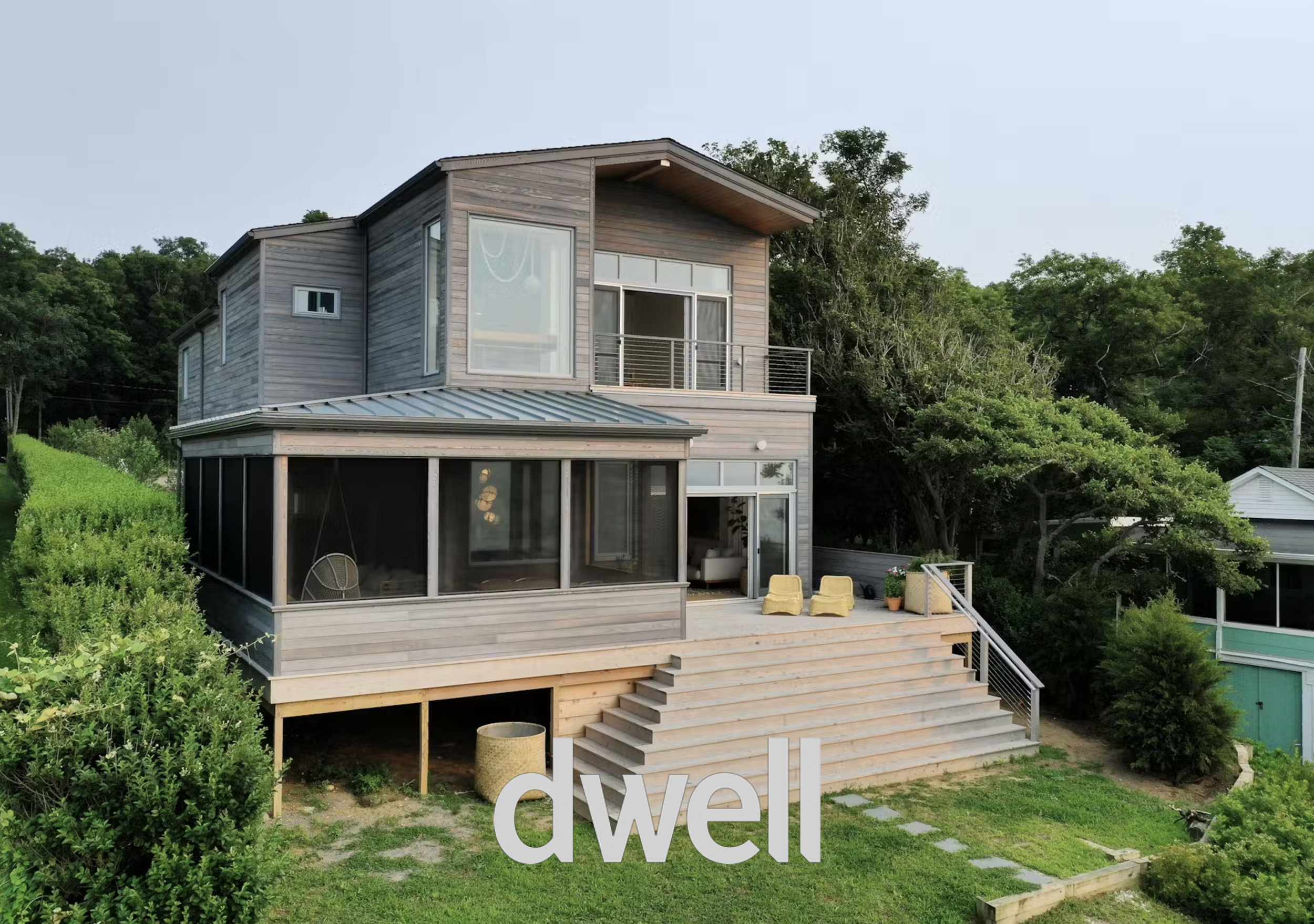 Modern multi-story house with large windows, wooden exterior, and a screened porch, surrounded by greenery and trees, with outdoor seating on a deck and steps leading down to a lawn.