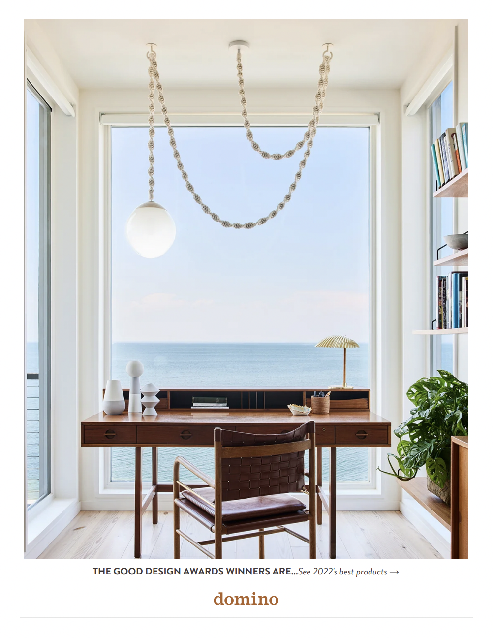 Interior office space with a wooden desk and a brown leather chair, large windows with ocean view, bookshelf on the right, plants, and decorative objects on the desk.