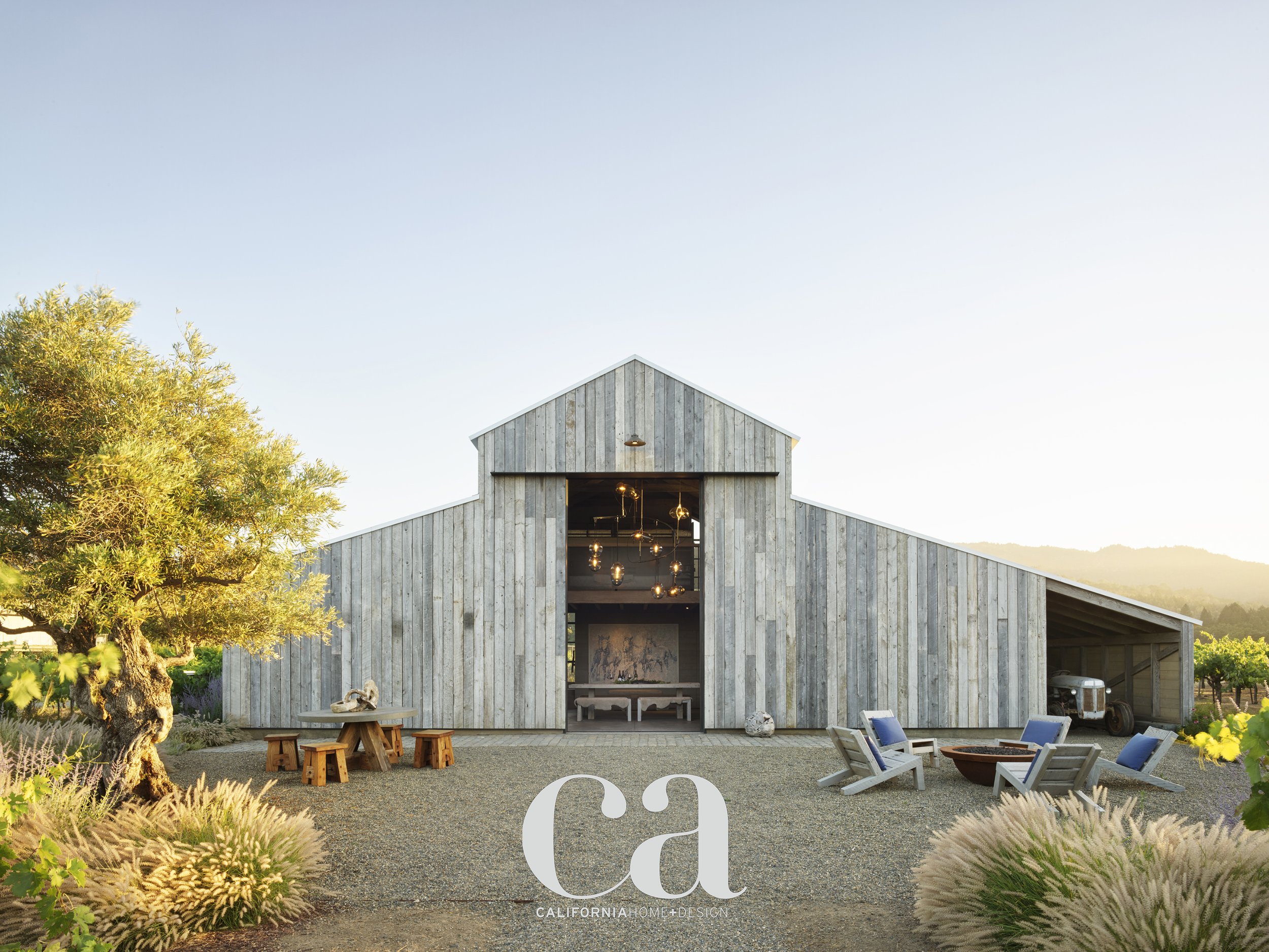A modern wooden barn with a large open entrance, outdoor seating area with Adirondack chairs, a table with benches, a vintage truck inside, and a tree with ornamental grasses in the foreground, under a clear sky.