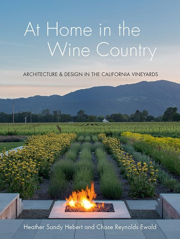 Book cover titled 'At Home in the Wine Country' with subtitle 'Architecture & Design in the California Vineyards,' showing a vineyard landscape with mountains, green rows of plants, and a modern fire feature in the foreground.