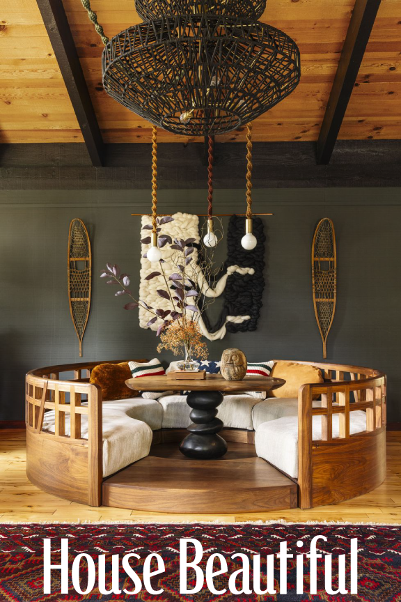 A cozy indoor space with a circular wooden seating area, a black pedestal table with decorative items, including a vase with dried plants, and artistic wall hangings on a gray wall, with a wooden ceiling overhead.