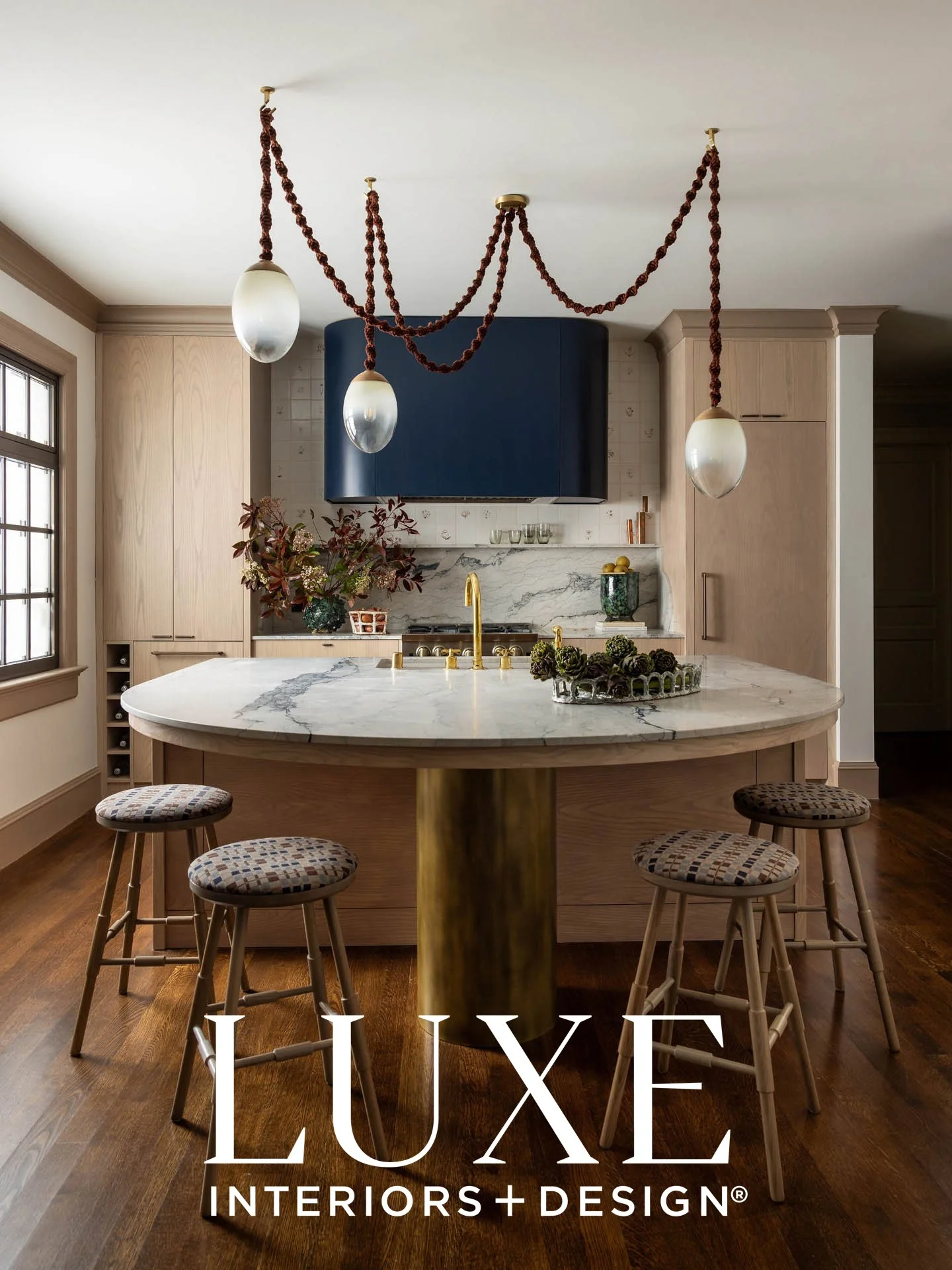 A modern kitchen with beige cabinetry, a marble island with a gold base, and a blue range hood. Hanging from the ceiling is a decorative Brightbound lighting fixture with Sunbrella Rope. The kitchen has hardwood floors and a window on the left side.