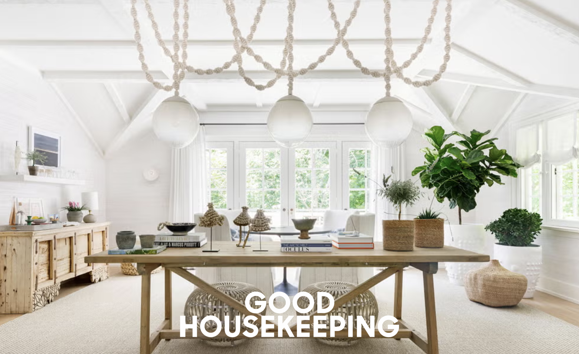 Bright and airy living room with white walls and ceiling, decorated with white curtains, green indoor plants, a large wooden dining table, and modern pendant lighting, with the overlay text "Good Housekeeping".