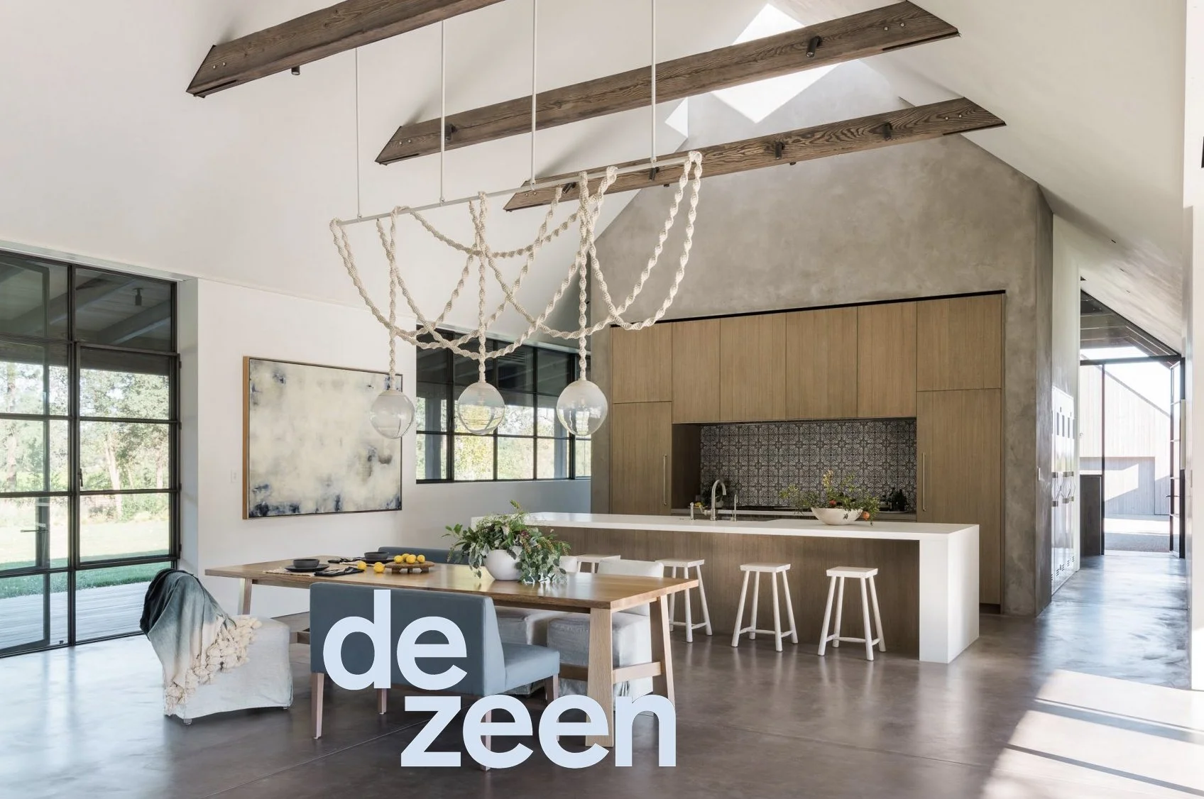 Modern open-concept kitchen and dining area with high ceilings, wooden beams, and large windows, featuring a counter with stools, a dining table with chairs, and decorative elements.