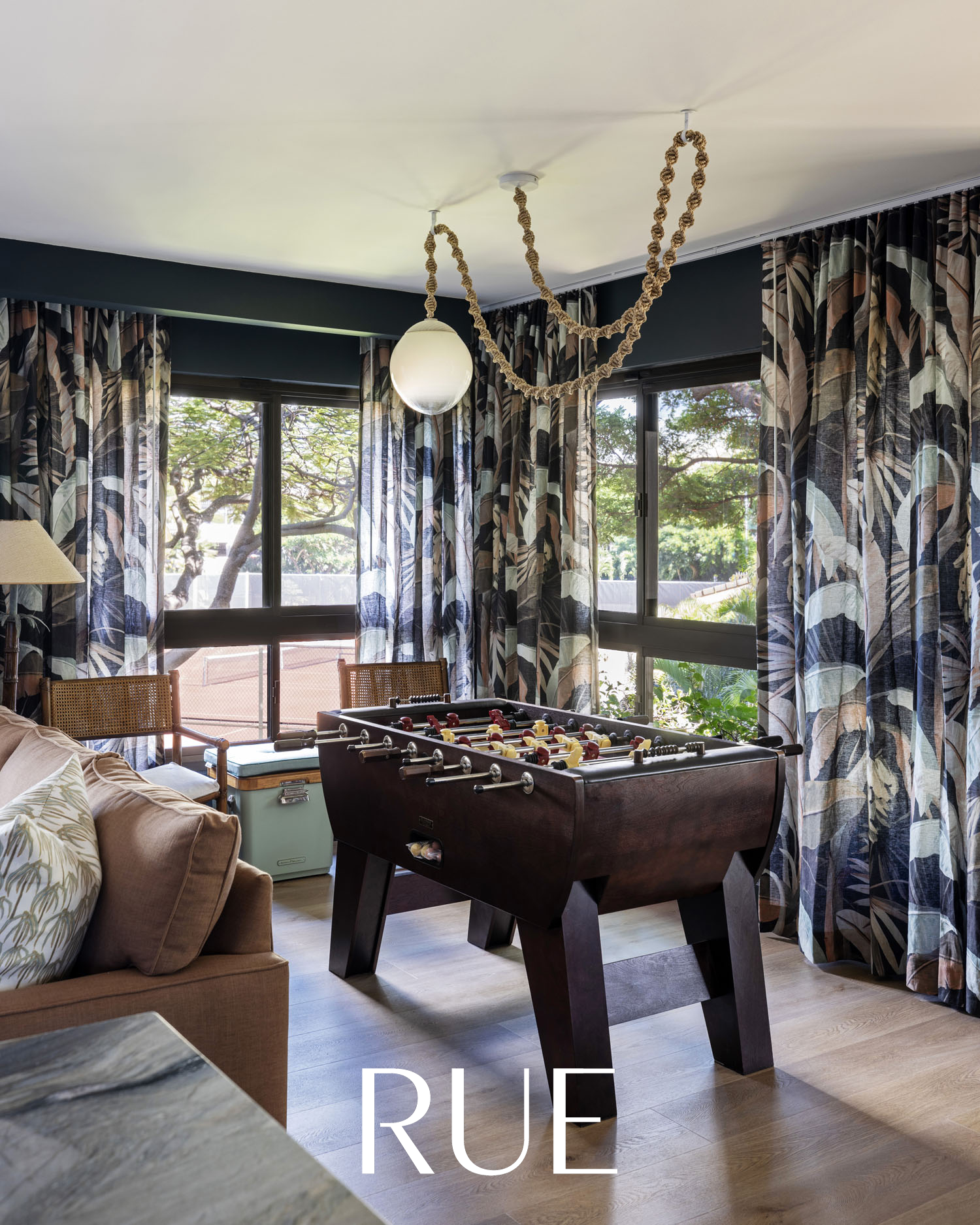 Interior of a living room with large windows, patterned curtains, a foosball table, a beige sofa with pillows, and greenery outside.