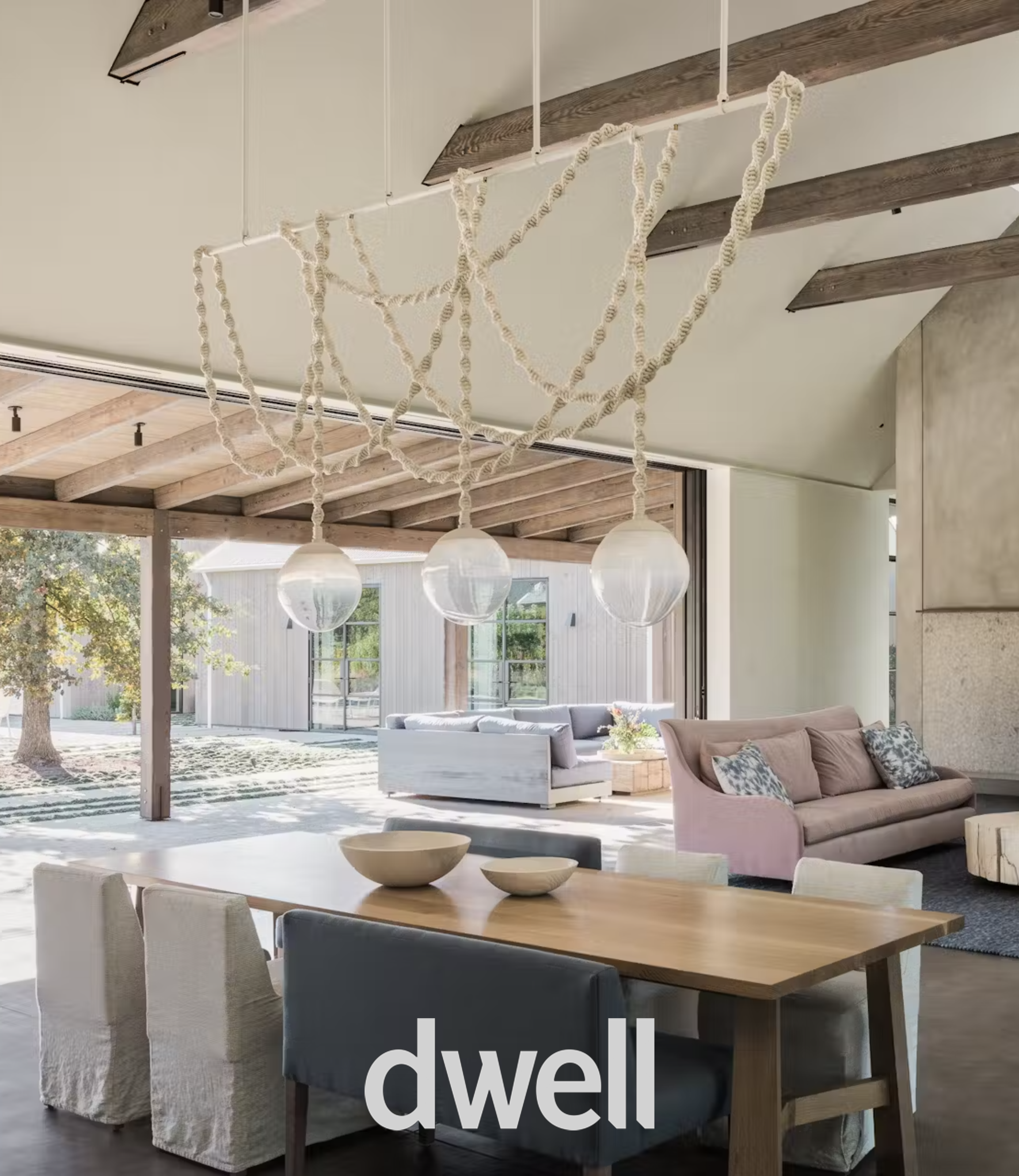 Open-concept living and dining area with modern furniture, hanging light fixtures, wooden beams, large windows, and outdoor patio visible through sliding doors.