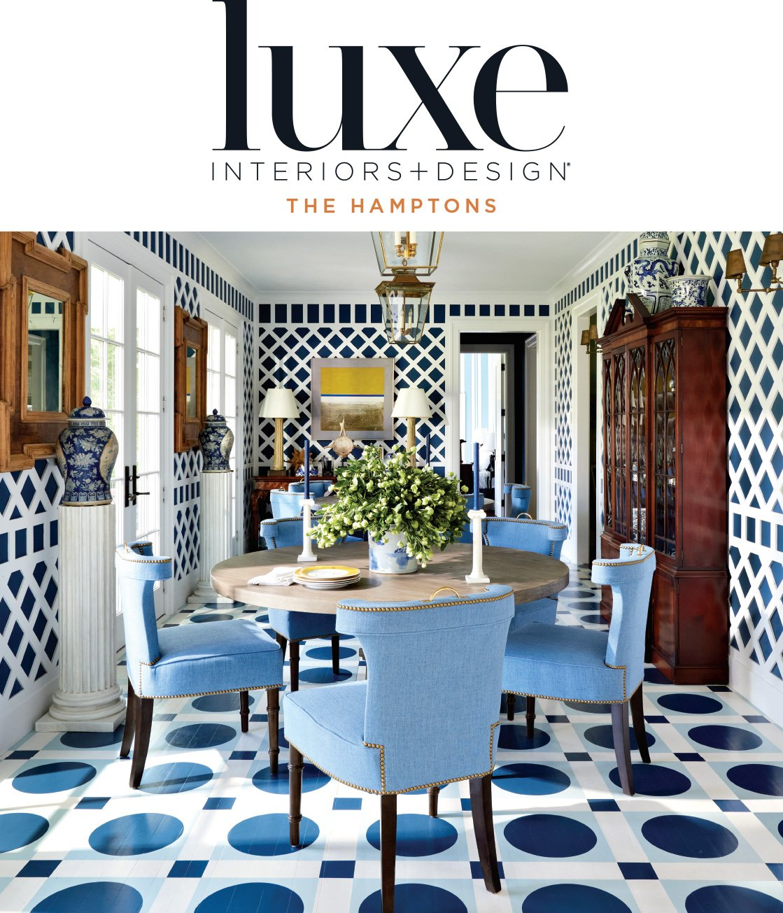 A dining room with blue and white patterned walls and floor, a round wooden table with a large floral centerpiece, six blue upholstered chairs, and wooden cabinets and decorative vases.