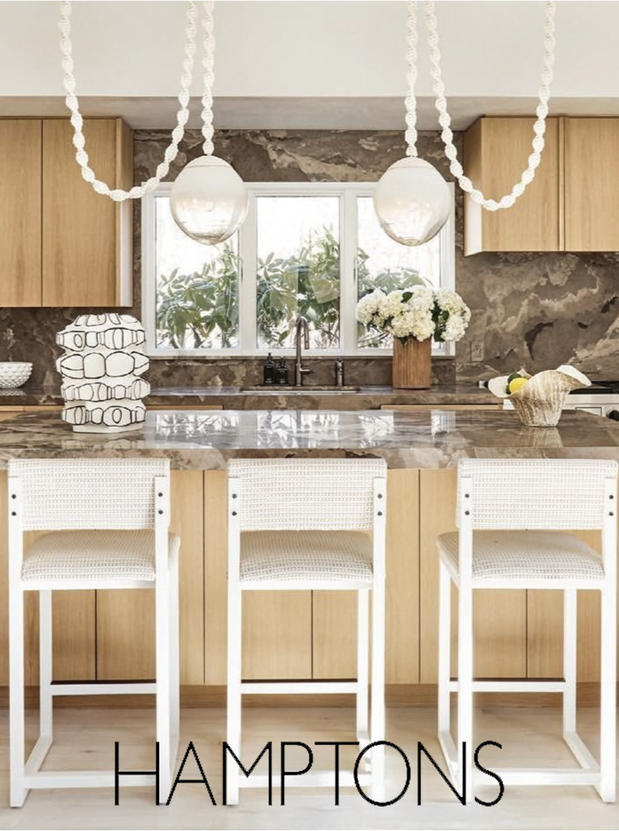 Modern kitchen with beige wooden cabinets, marble countertop, and two white barstools, decorated with a large glass vase of white flowers, a basket with fruit, and paper lamp shades, with large window and greenery outside.