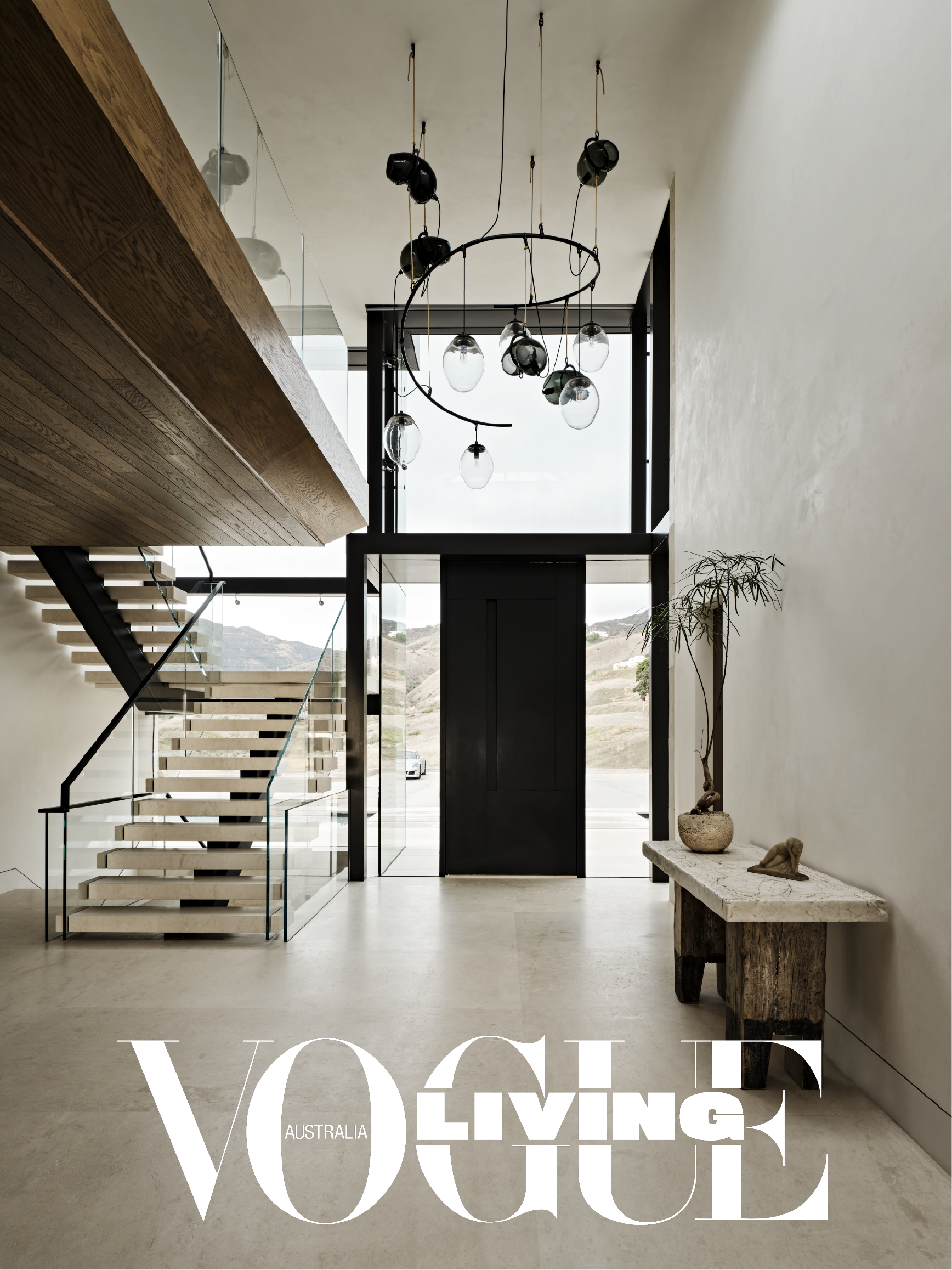 Modern interior entrance hall with a minimalist design, featuring a staircase with wooden steps and glass railing, hanging pendant lights, large front doors, a potted plant, and a rustic wooden bench.