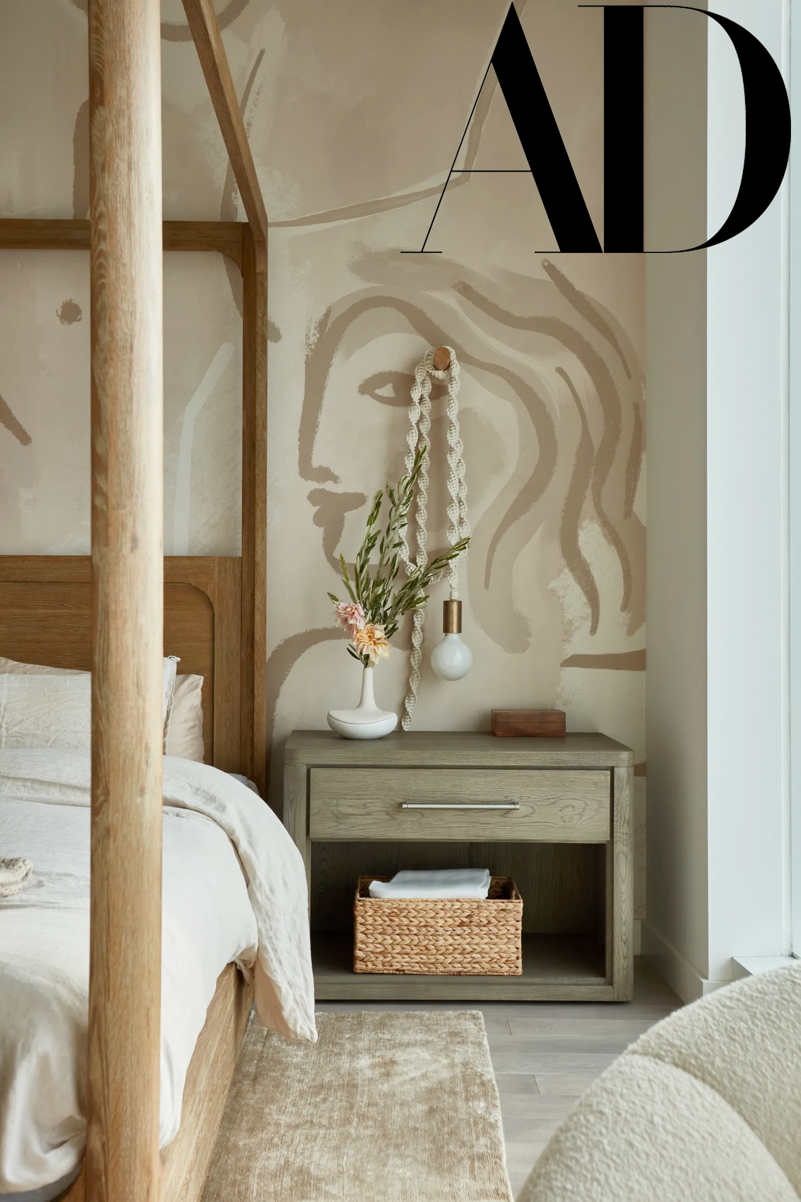 A bedroom corner with a wooden bed and a nightstand. The wall has a painted outline of a woman's face and hair. The nightstand holds a vase with flowers, a hanging braid, a light bulb, and a small box. There is a woven basket with towels below the ni