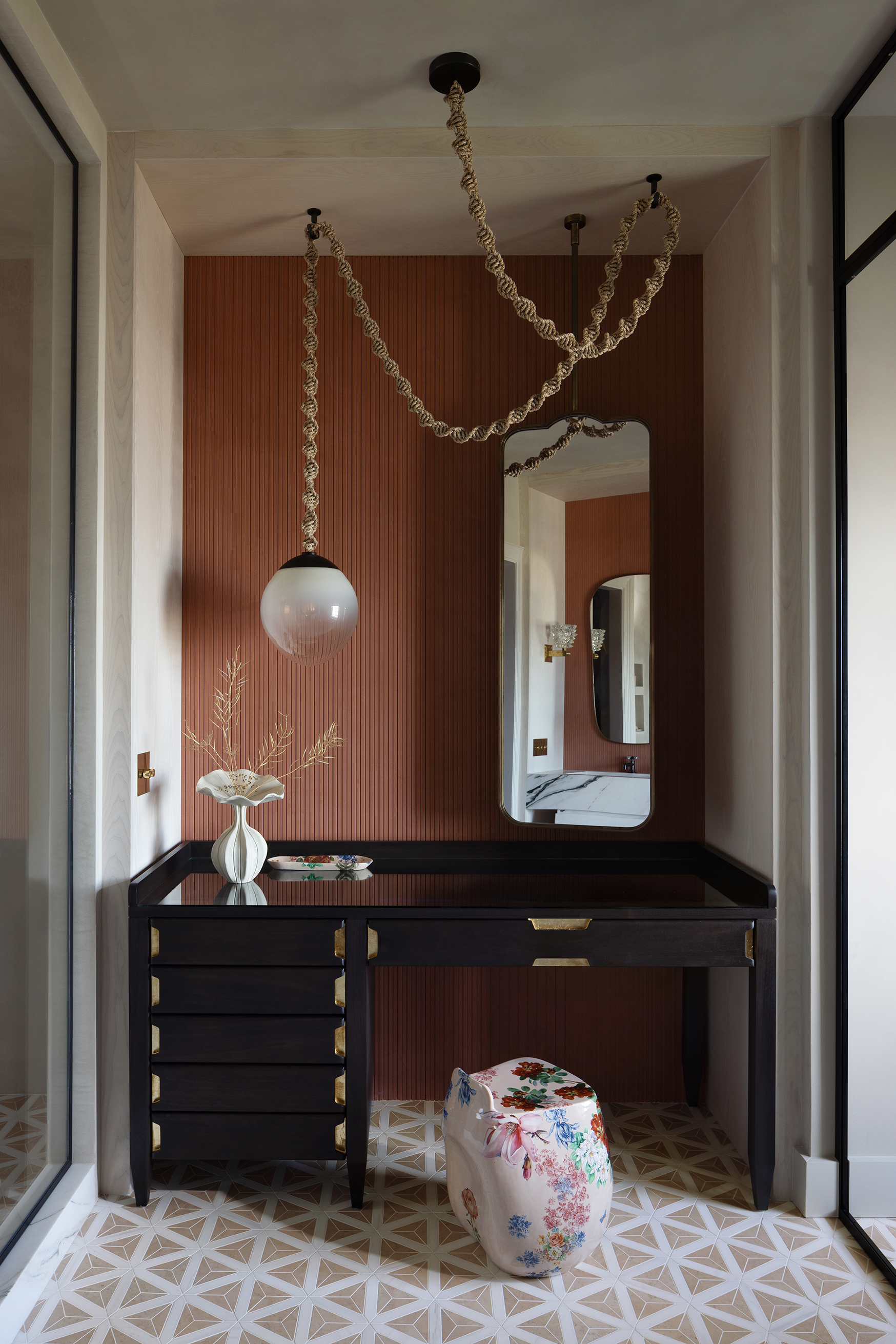 Modern entryway with a black console table against a rust-colored textured wall, a decorative white vase with branches, a floral upholstered stool, and a large mirror reflecting the bathroom and lighting fixture.