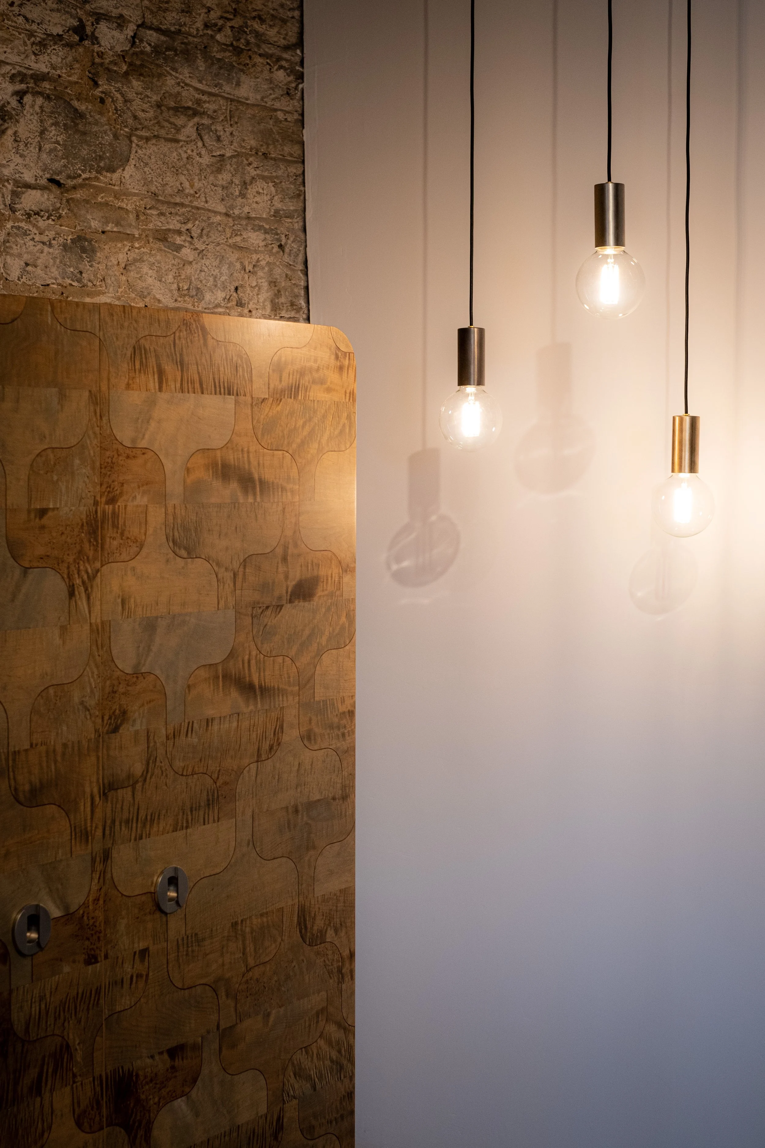 Three hanging light bulbs with exposed filaments casting shadows on a white wall next to a stone wall and a wooden decorative panel.