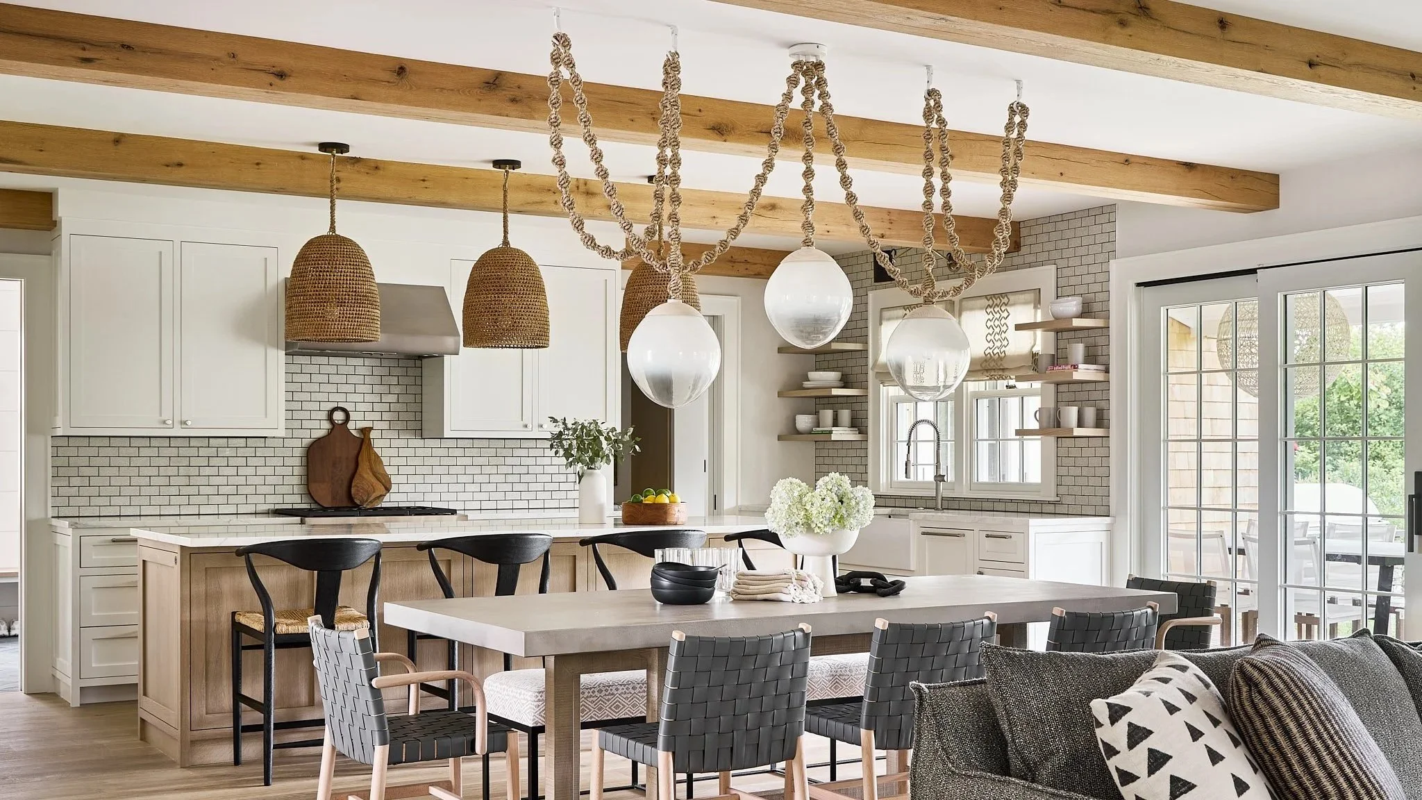 A modern kitchen interior with a large dining table, black woven chairs, white cabinetry, open shelves, a white tiled backsplash, a window, sliding glass door, hanging pendant lights, and decorative rope accents on the ceiling.
