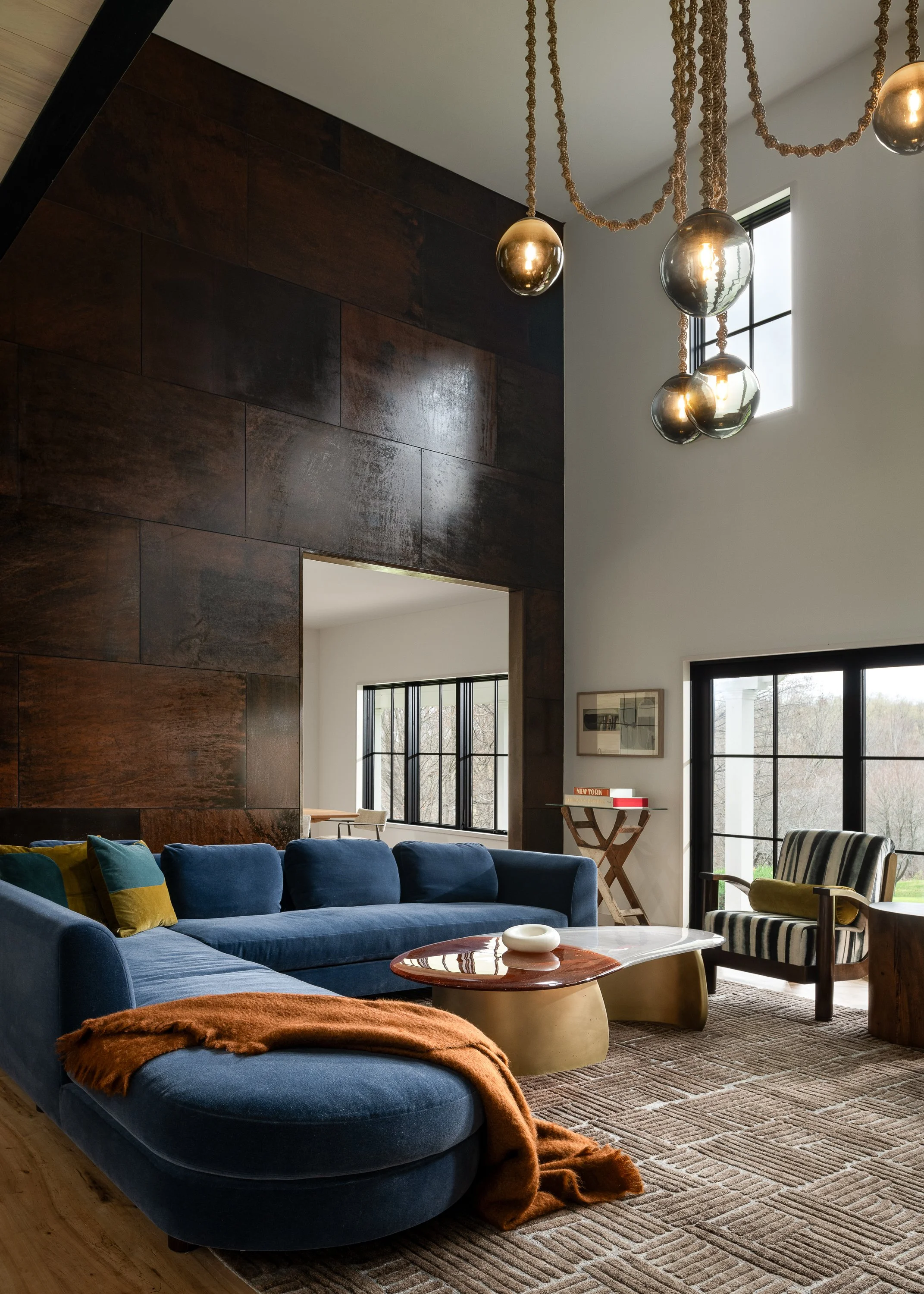 Living room with a large blue sectional sofa, striped armchair, modern pendulum chandelier, dark wood accent wall, and large windows with black frames in a contemporary style.