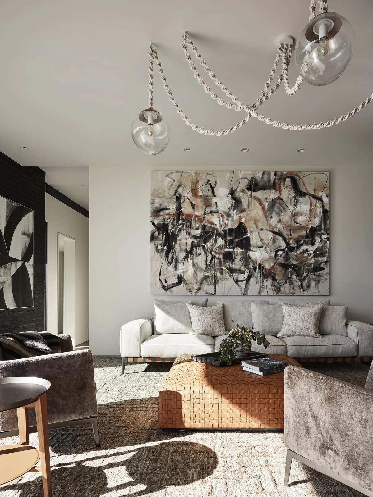 Modern living room with a white sofa, abstract wall art, orange woven ottoman, and velvet chairs, with sunlight casting shadows on the beige carpet.