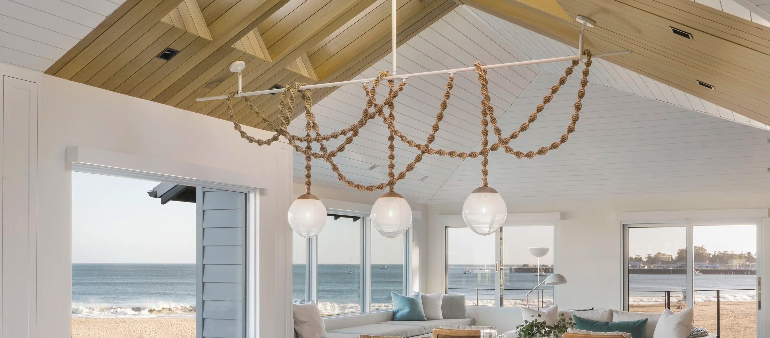 Beachside living room with panoramic ocean view, white walls, large windows, a cozy white sofa with pillows, and a hanging light fixture decorated with rope.