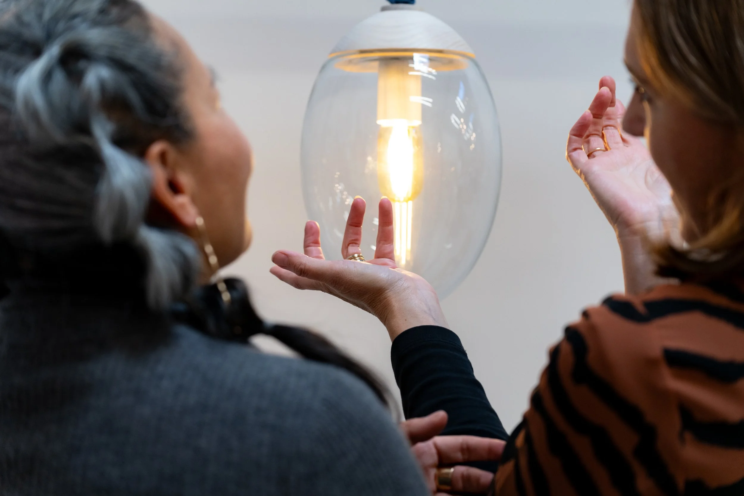 Two women are standing near a wall-mounted lamp, with one lady holding her hand in front of her face and the other lady gesturing toward the lamp.