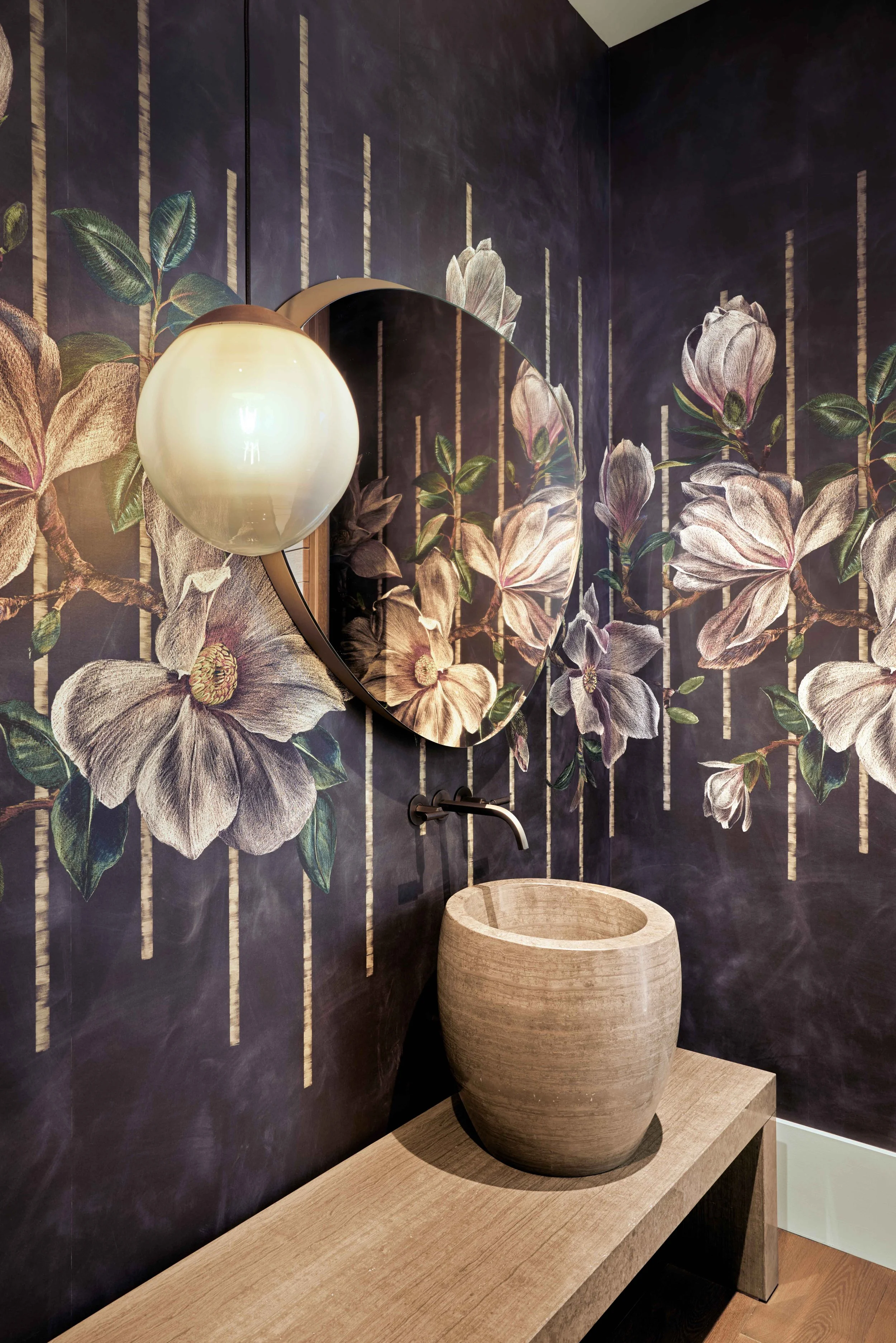 A bathroom with dark floral mural, round mirror, Brightbound Sila studio-blown pendant lighting, and stone sink on a wooden counter.