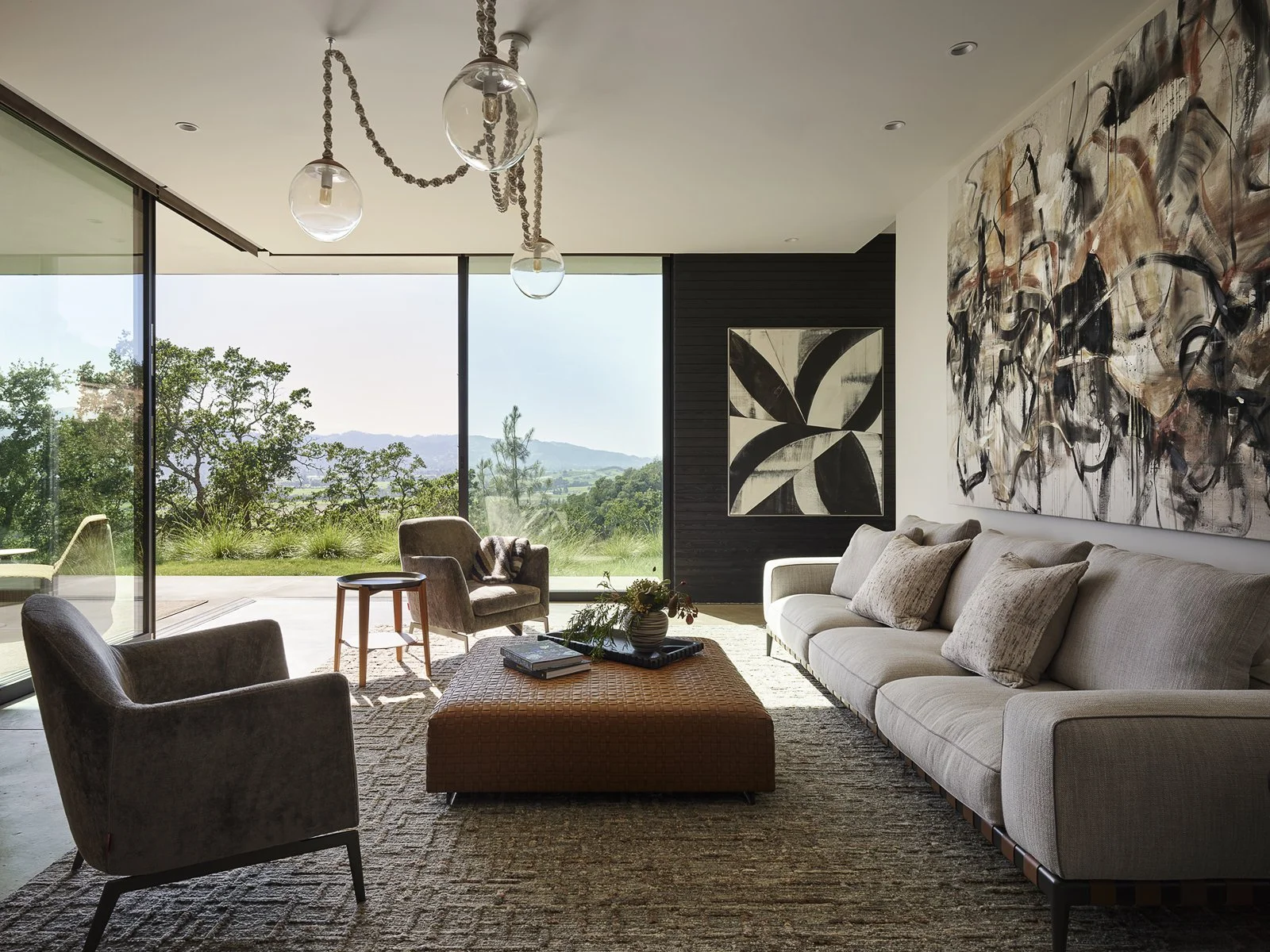 Modern living room with large windows showing landscape, beige sofa, armchairs, a woven ottoman, contemporary art, and hanging glass light fixtures.