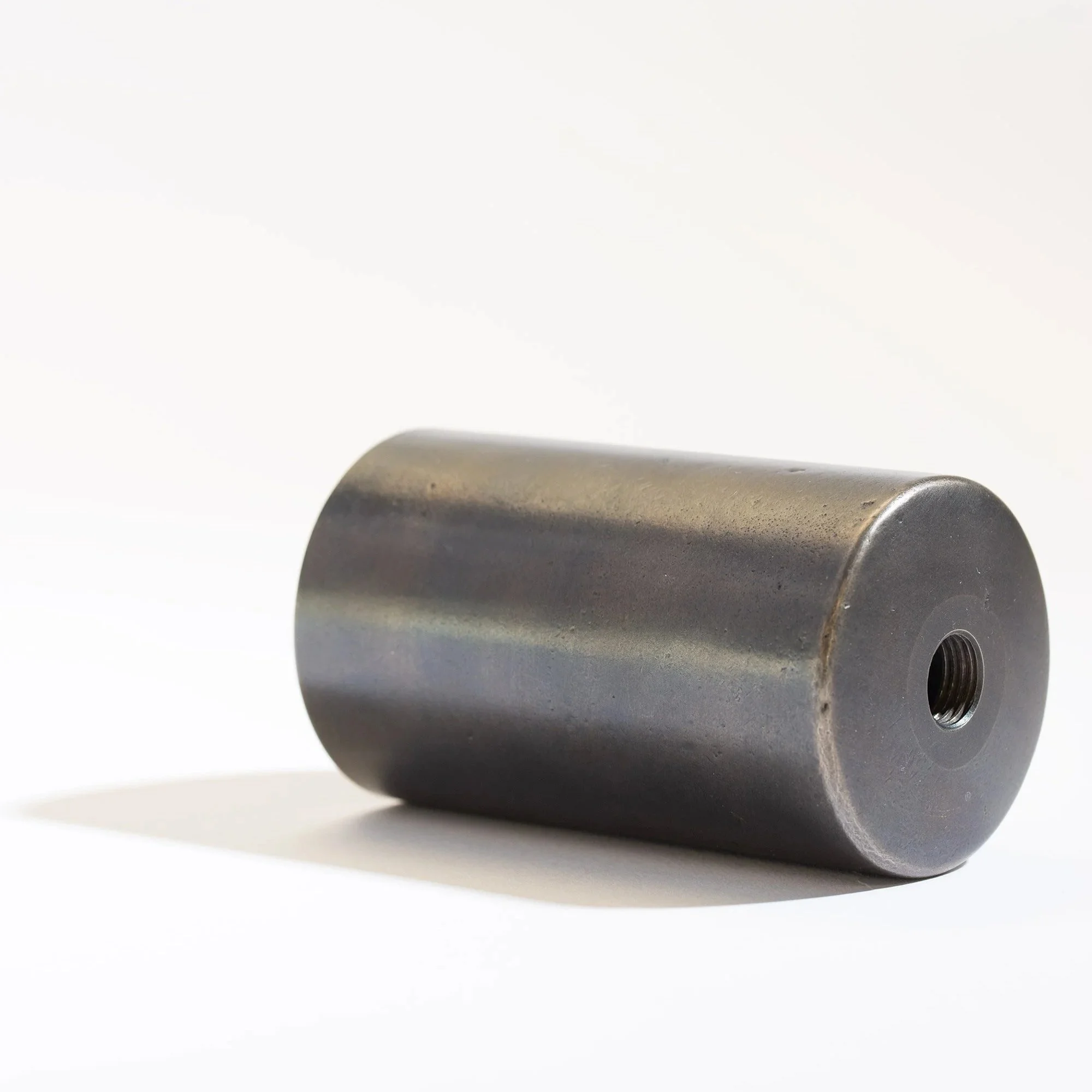 A metal cylindrical object with a threaded hole at one end, placed on a white surface with shadows underneath.