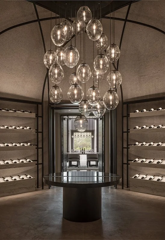 Elegant wine cellar with Gemma studio-blown glass lighting chandelier, a round reflective table in the center, and shelves filled with wine bottles on either side, with a seating area visible through a doorway in the background.