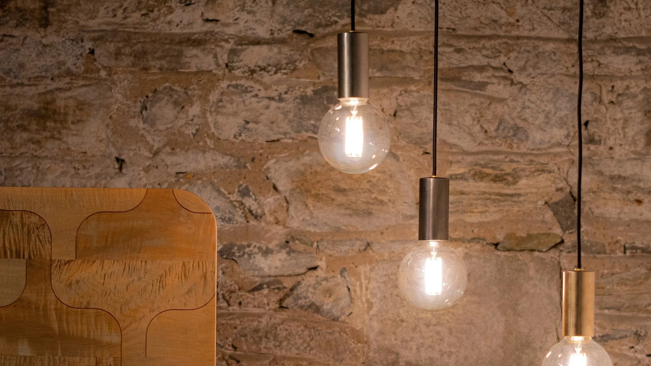 Three hanging pendant lights with exposed bulbs above a wooden piece of furniture in front of a stone wall.