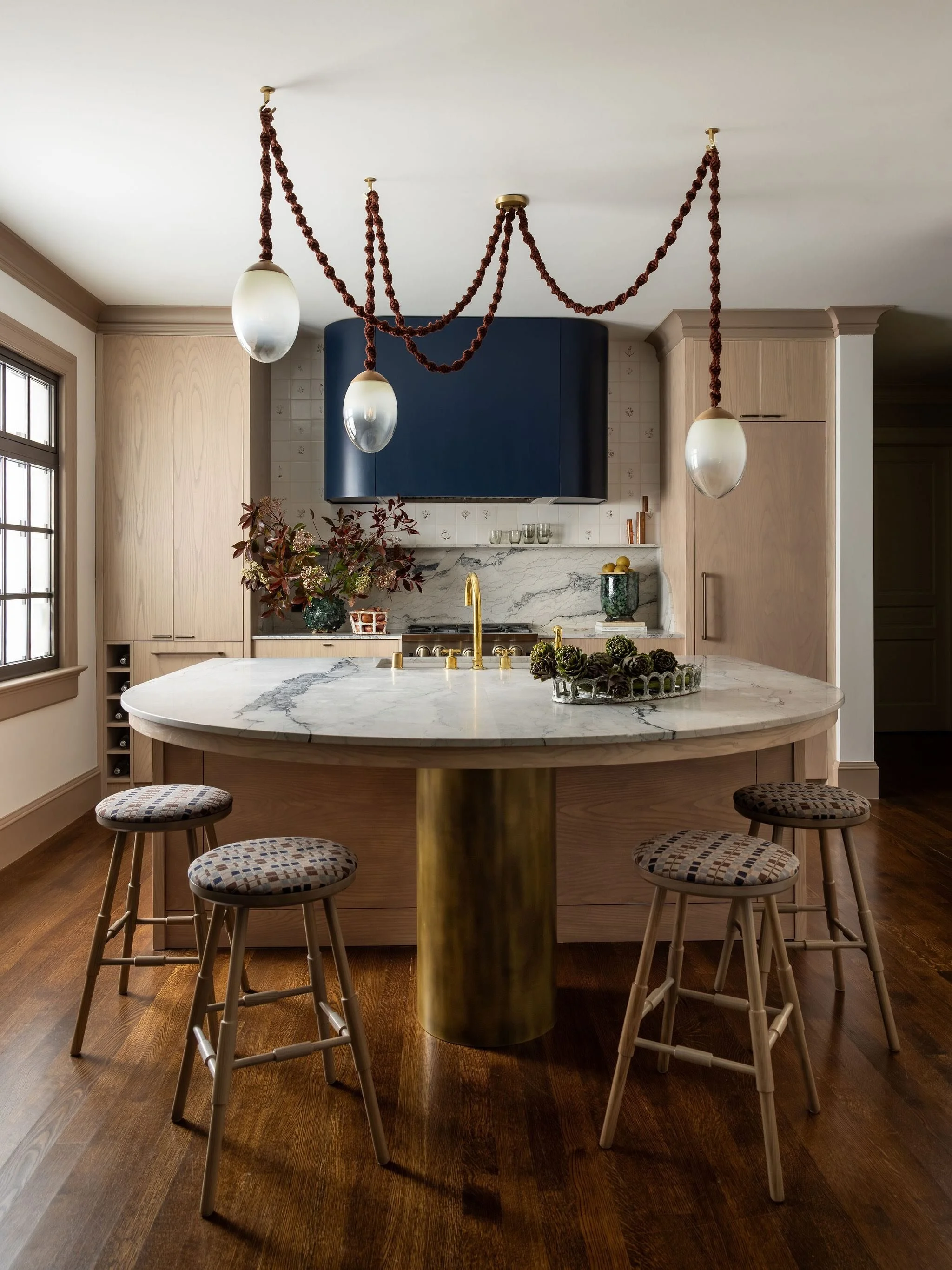 Modern kitchen with a large marble island, gold accents, Olive Helix studio-blown glass and Sunbrella Rope chandelier lighting, beige cabinetry, and a blue range hood.