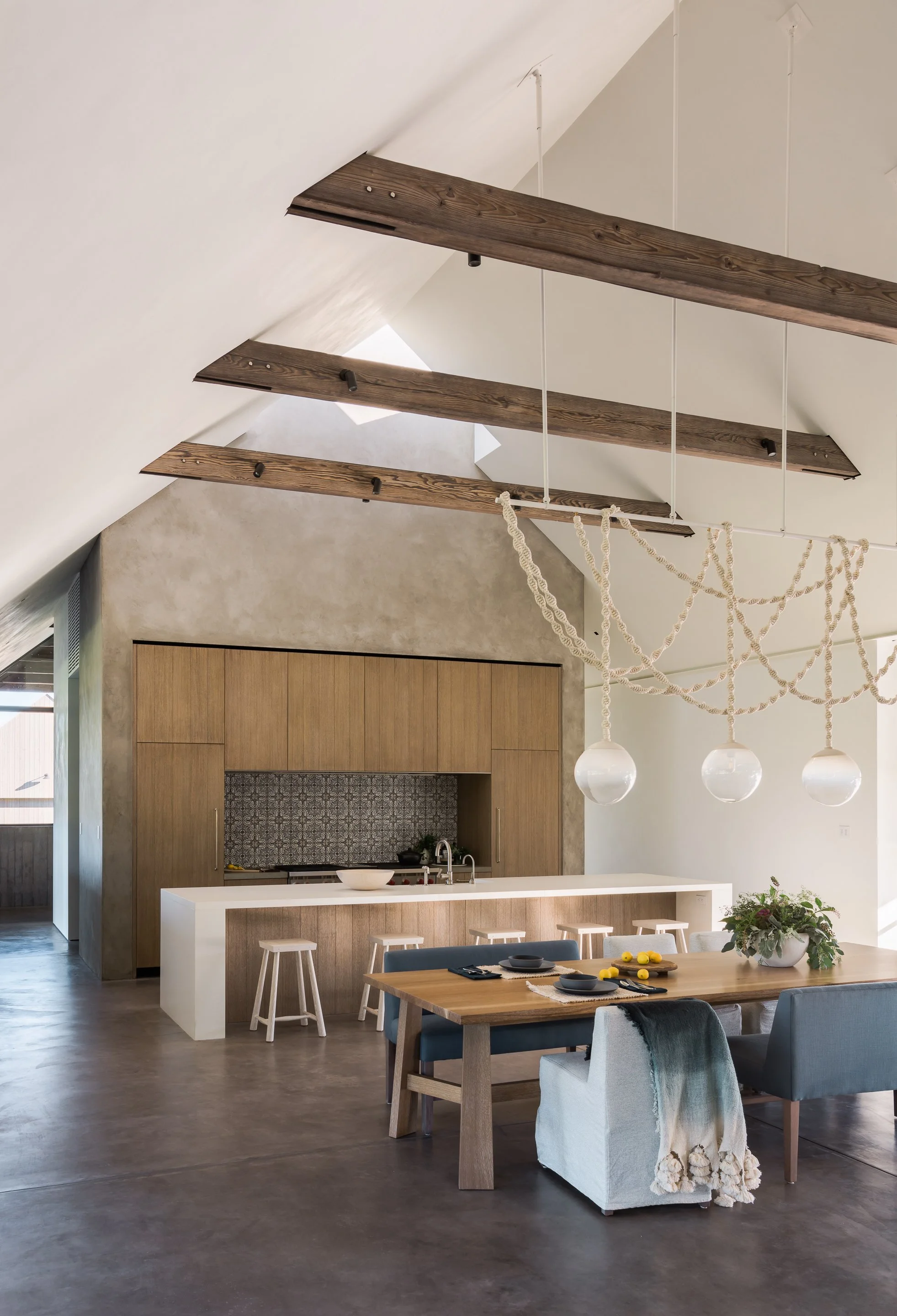 Modern kitchen and dining area with vaulted ceiling, exposed wooden beams, and minimalist decor. The space features a built-in wooden cabinet, a kitchen island with a sink, and a wooden dining table with blue and white chairs, decorated with tableware, lemons, and a potted plant.