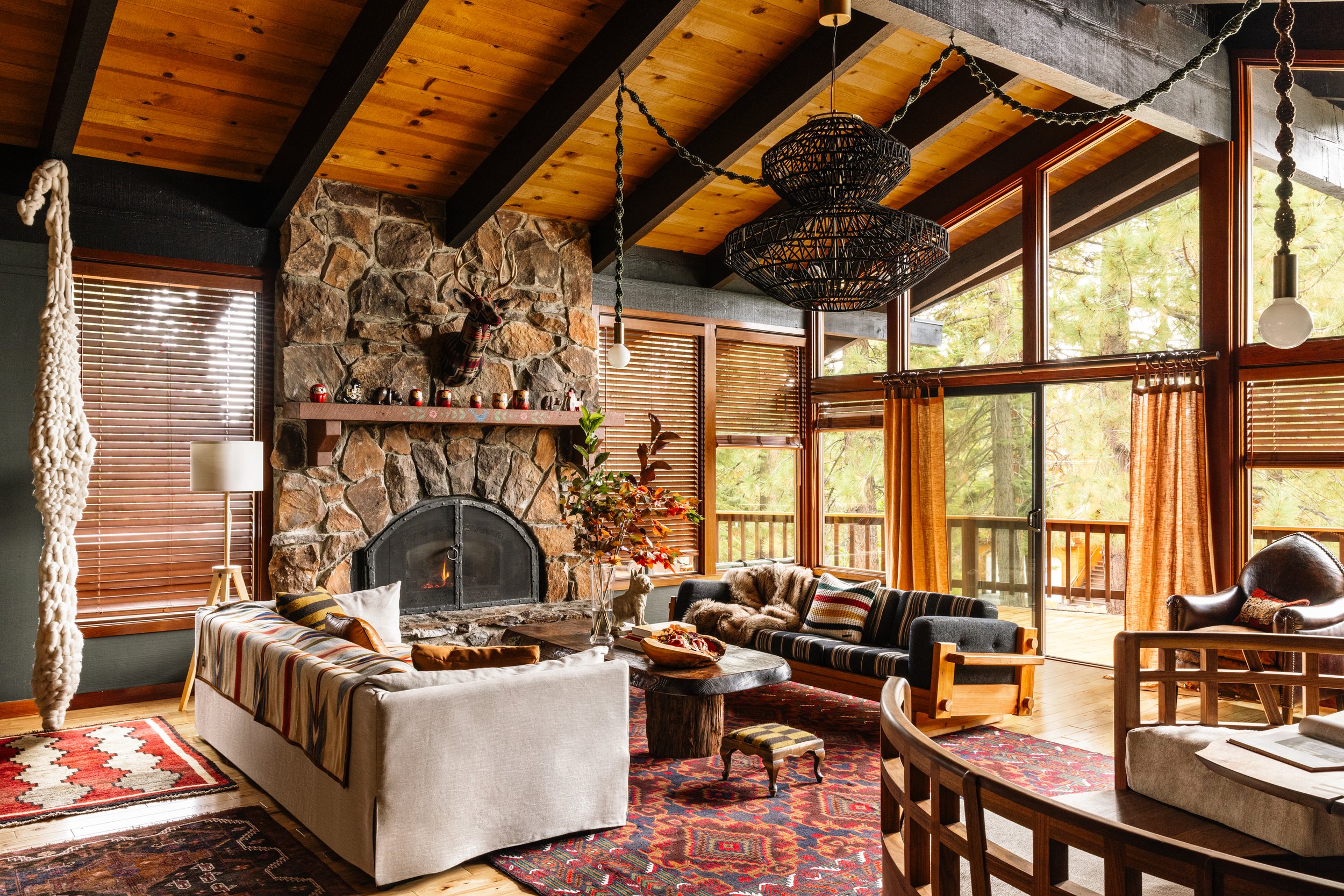 Living room with wooden ceiling, stone fireplace with mounted deer head, large windows, and furniture including sofas, chairs, and a coffee table with fall-themed decor.