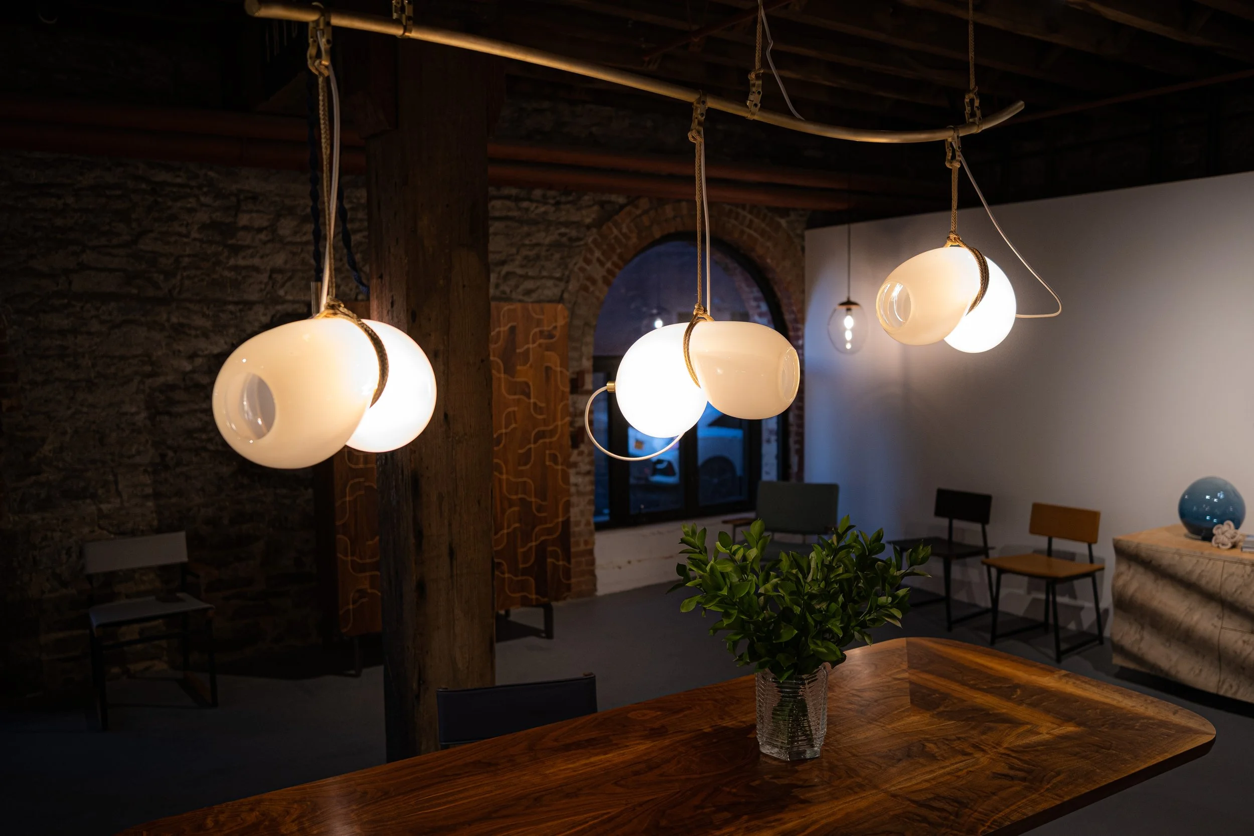 Modern interior room with hanging white spherical pendant lights, a wooden table with a green plant in a glass vase, chairs, and decor items against an exposed brick wall.