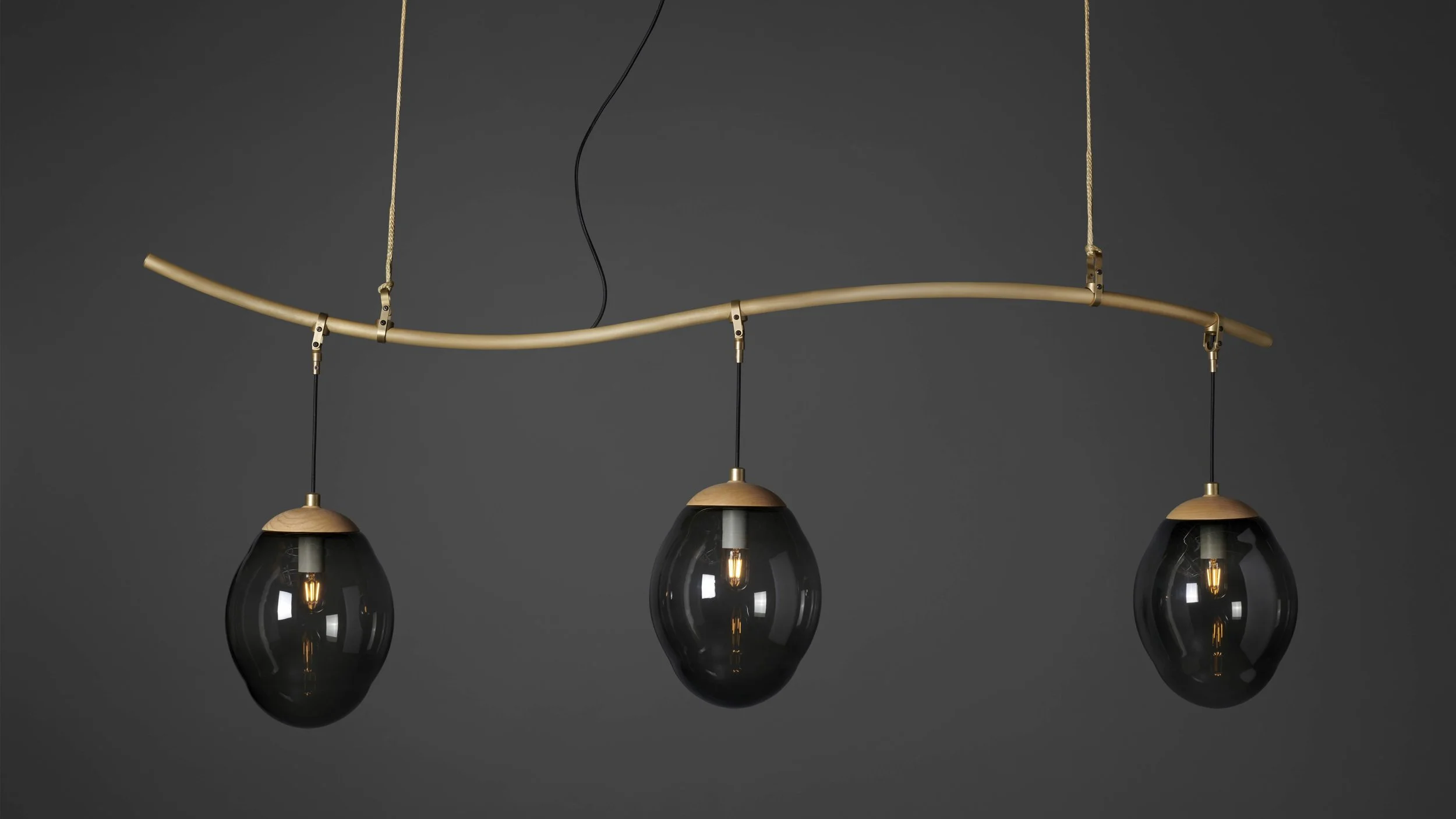 Modern pendant light fixture with three black glass bulbs hanging from a curved wooden and gold-colored track against a dark background.