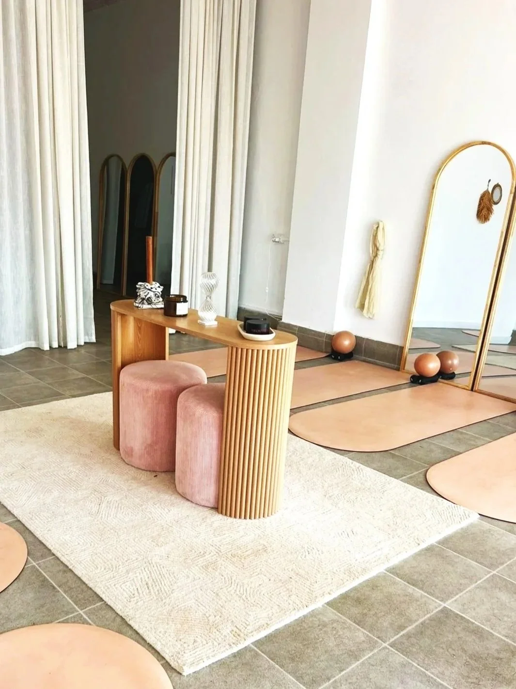 A minimalist interior space with a small wooden table, pink velvet ottomans, a large mirror with a gold frame, and a light-colored rug on a tiled floor.