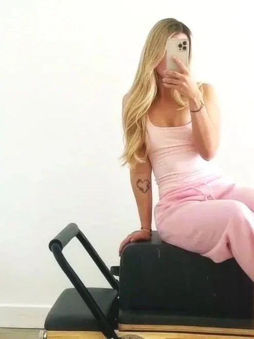 Woman with long blonde hair sitting on an exercise bench, taking a mirror selfie with a smartphone, wearing a light pink outfit, in a room with white walls.
