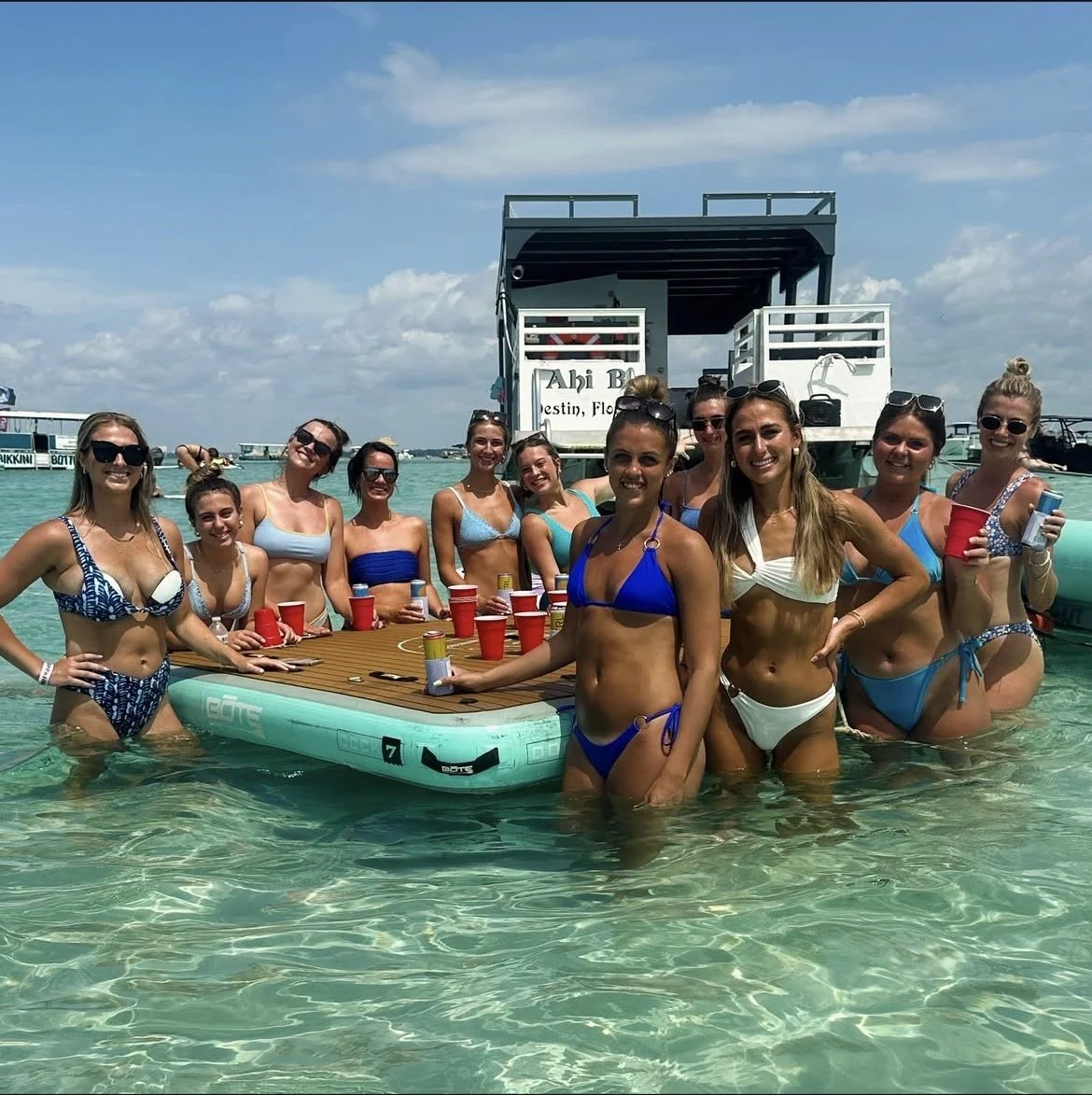 Group of women in swimsuits standing in shallow water around a floating platform with red cups, boats in the background, and a partly cloudy sky.