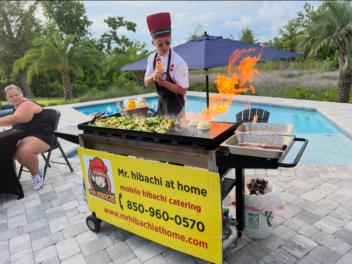 A chef, wearing sunglasses and a tall red hat, grills food with a flame on a patio near a swimming pool. The chef is making hibachi-style dishes, with food cooking on a flat top grill. A woman is seated at a table nearby. There is a yellow sign on the grill advertising Mr. Hibachi at home catering.