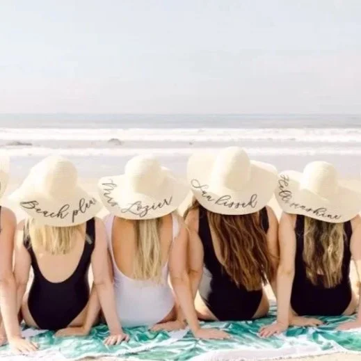 Five women sitting on a beach with their backs to the camera, wearing large sun hats with various words written on them, enjoying a sunny day by the ocean.